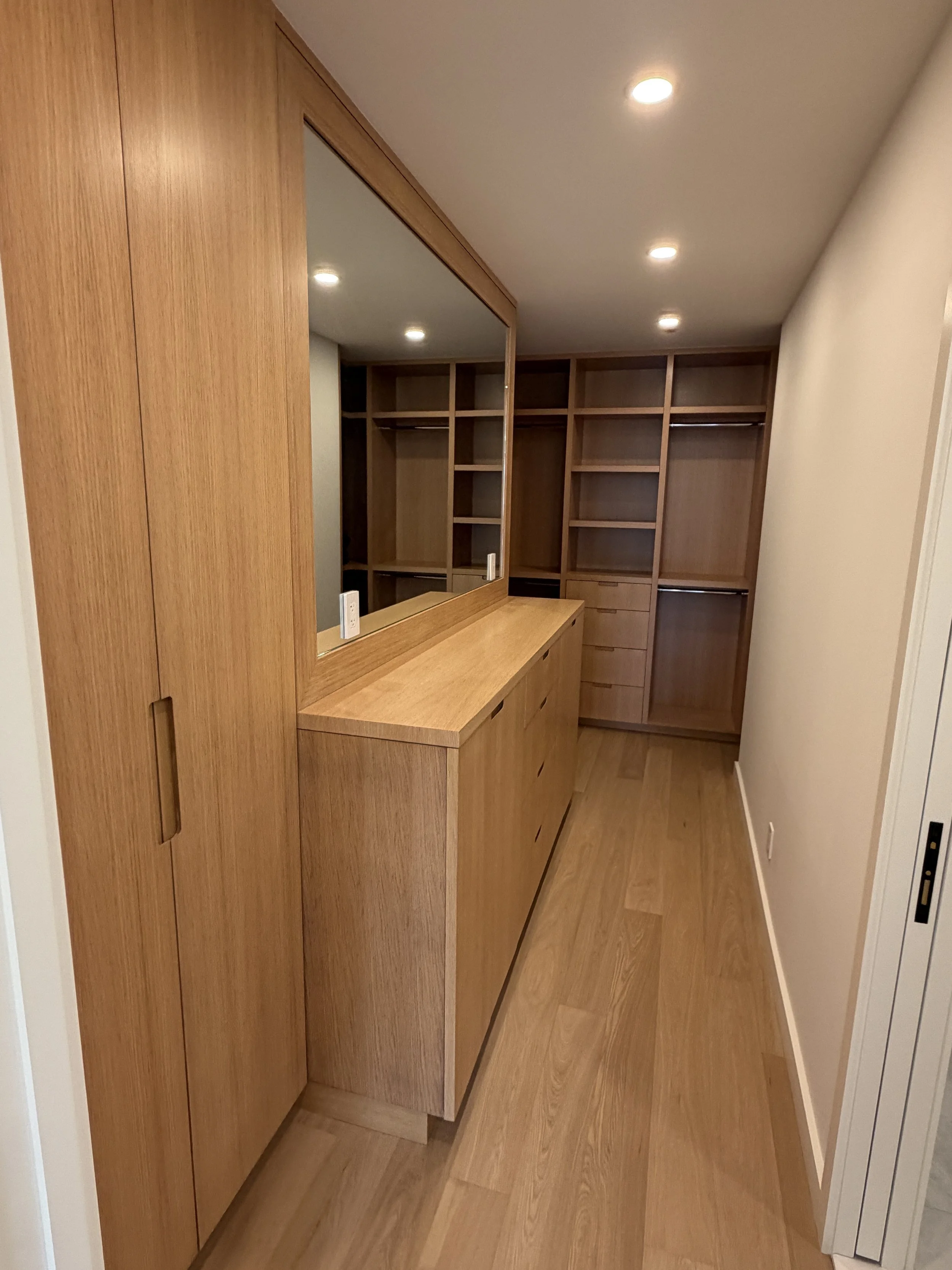Empty walk-in closet with built-in wooden shelves, drawers, and sliding mirror doors, illuminated by ceiling lights.