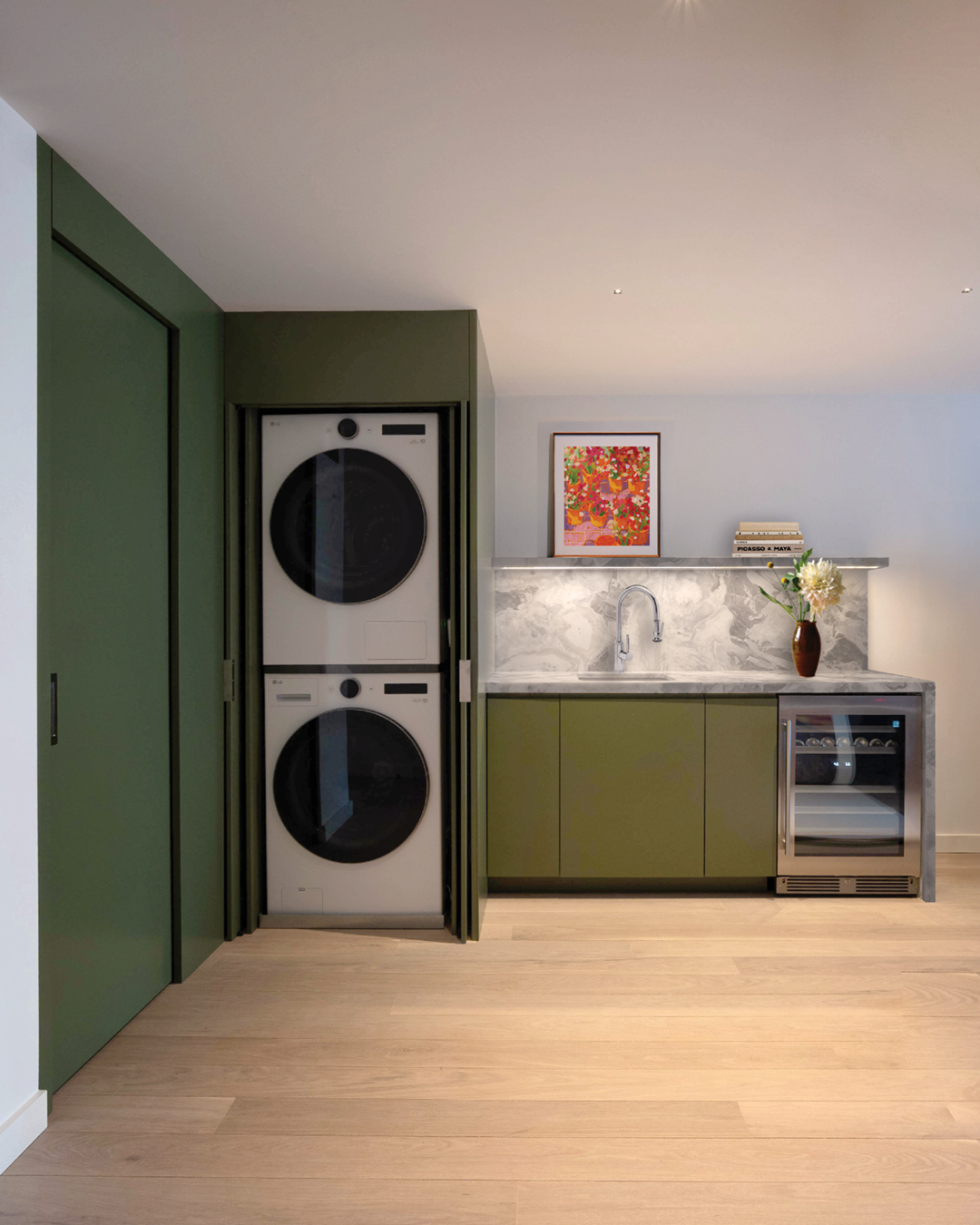Laundry area with stacked washer and dryer, green cabinets, marble countertop with sink, artwork, books, flower vase, and small wine fridge on light wood flooring.