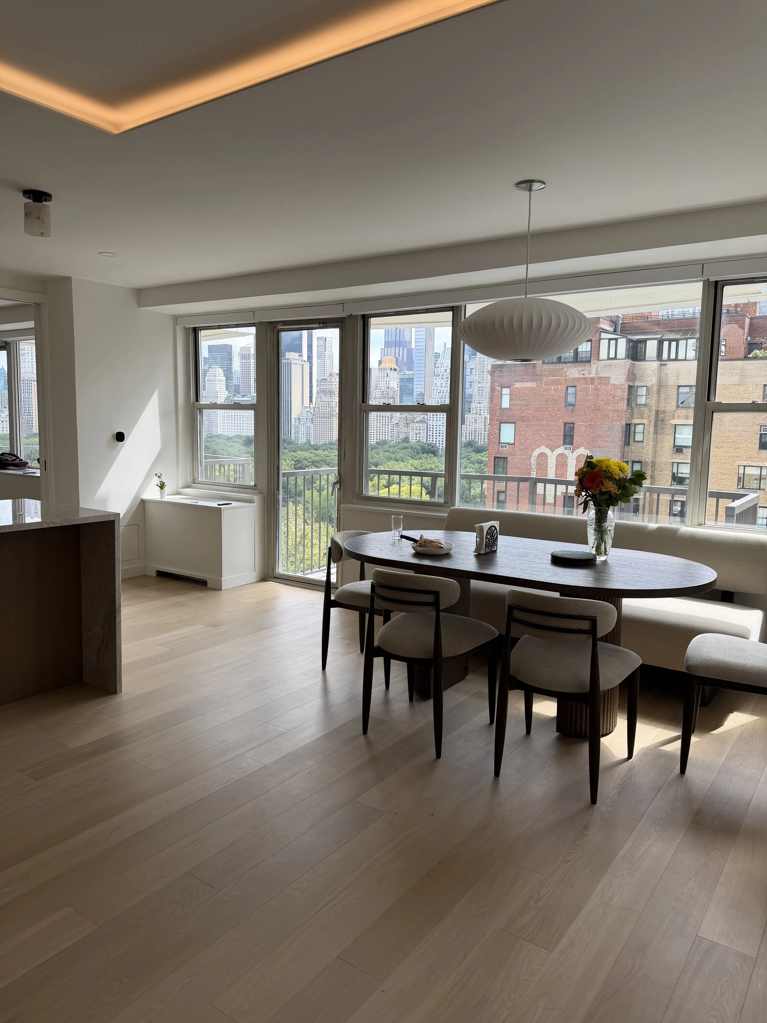 Modern dining room with large windows showing city buildings and greenery outside. Features include a round dining table with chairs, a vase of flowers, and a pendant light fixture.