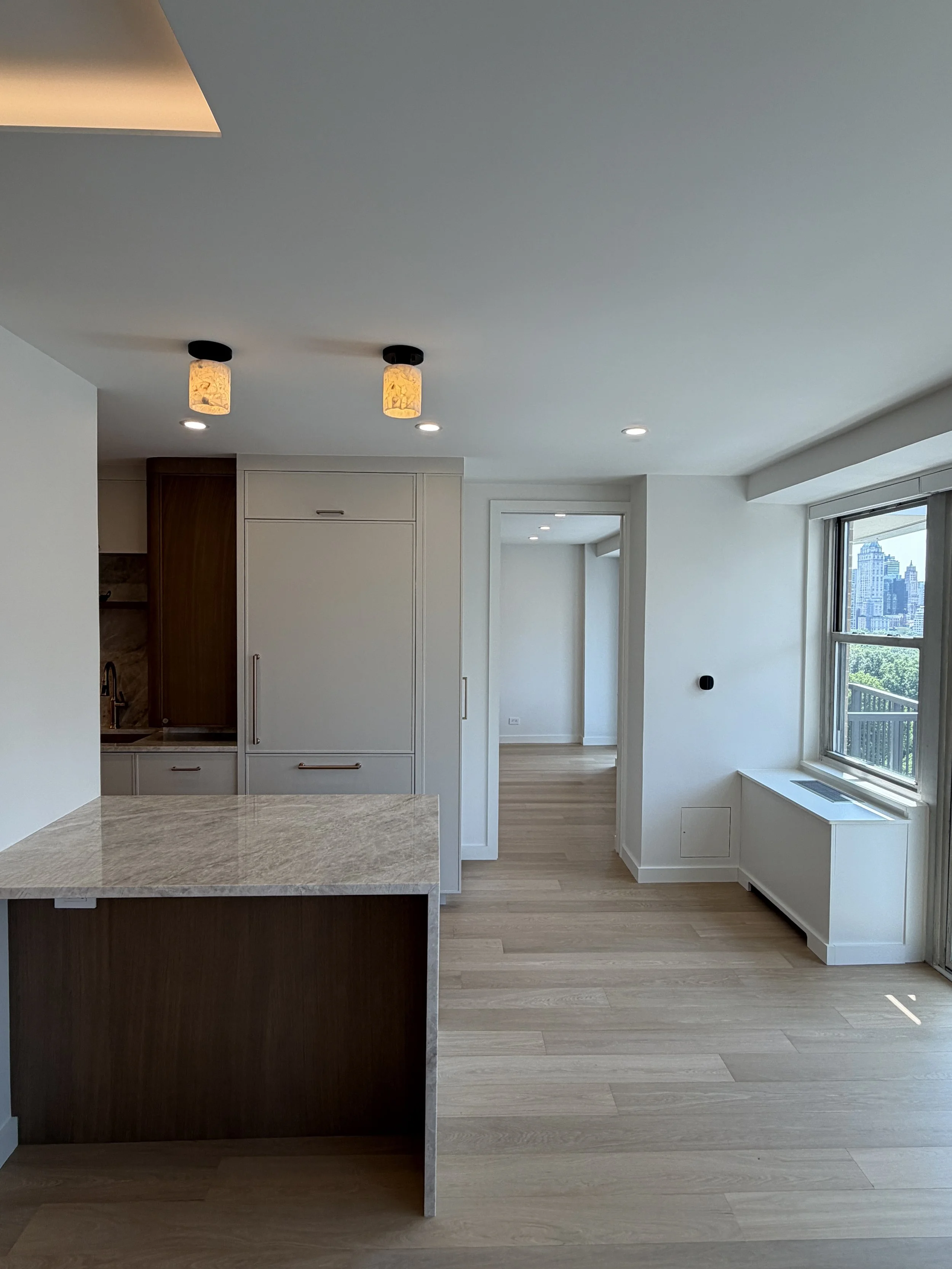 A modern, minimalistic kitchen and living area with light wooden flooring, white walls, and large windows showing a city skyline. Recessed lighting and two pendant lights are on the ceiling. A corridor leads to another room.