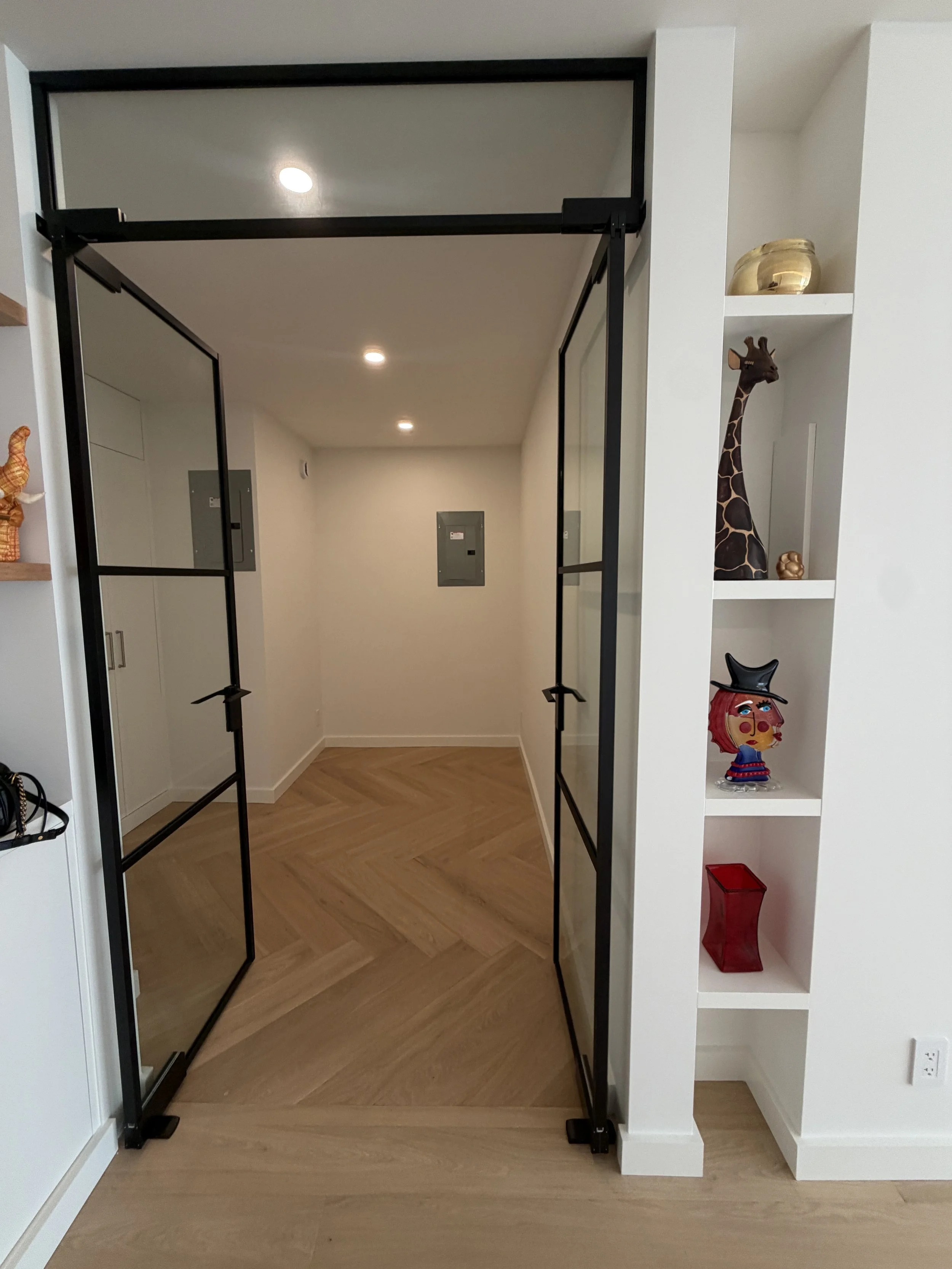 A black metal and glass sliding door in the process of being installed in a hallway, with a white wall and built-in shelves decorated with a giraffe sculpture, a gold bowl, a colorful abstract figure, and a red vase.