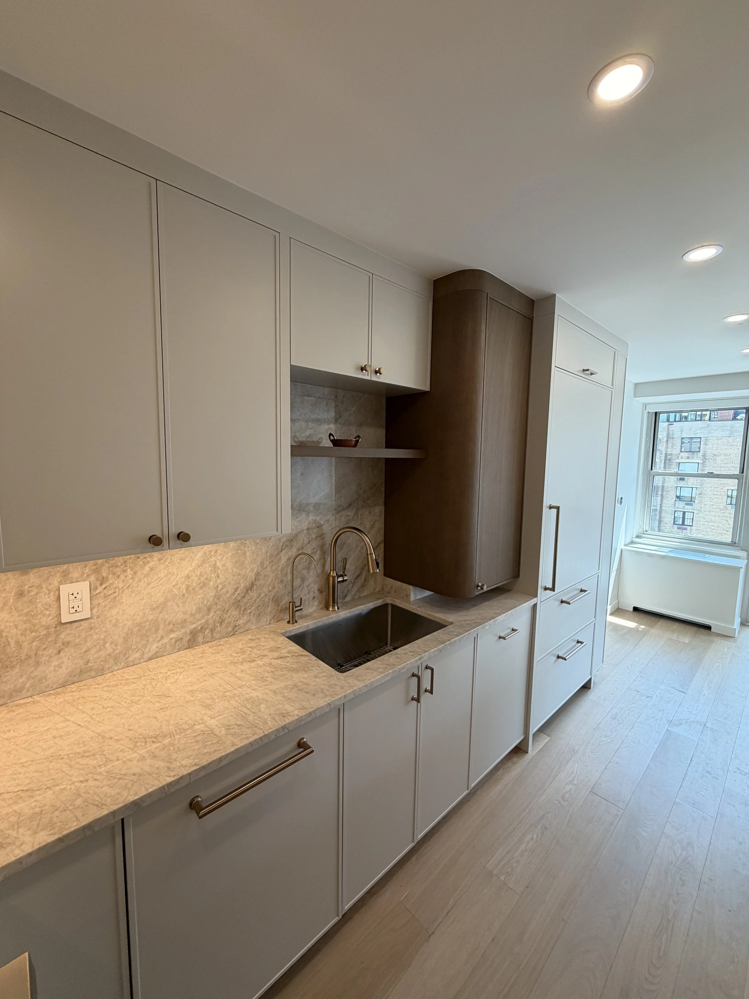Modern kitchen with beige and wooden cabinets, marble countertop, stainless steel sink, and a window with city view.