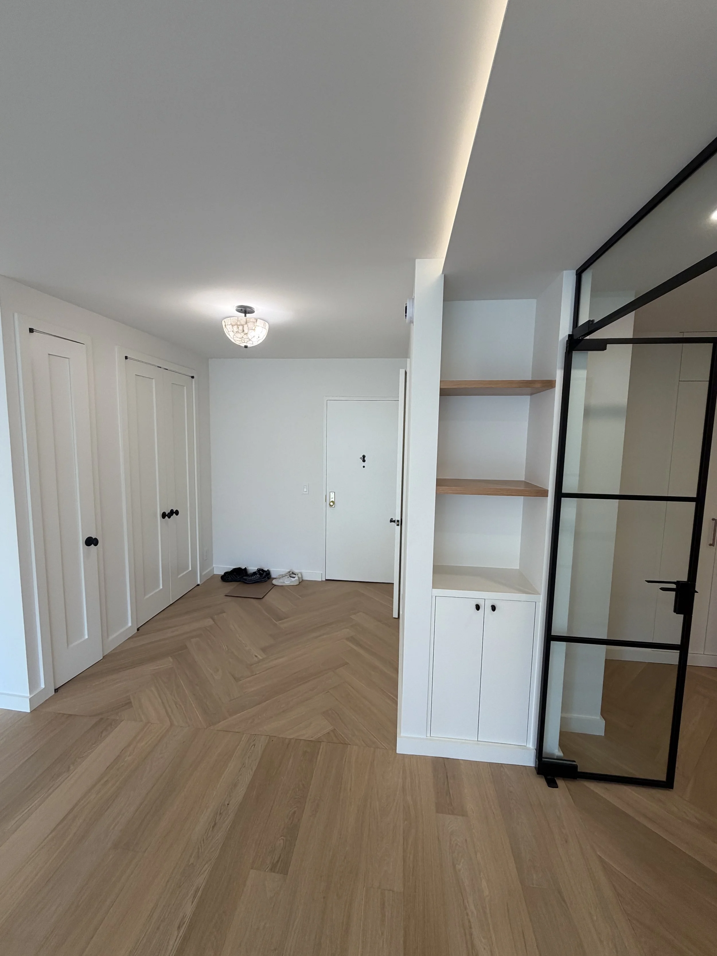 A modern interior space with white walls, wooden flooring arranged in a herringbone pattern, built-in white cabinets, and a glass and metal sliding door.