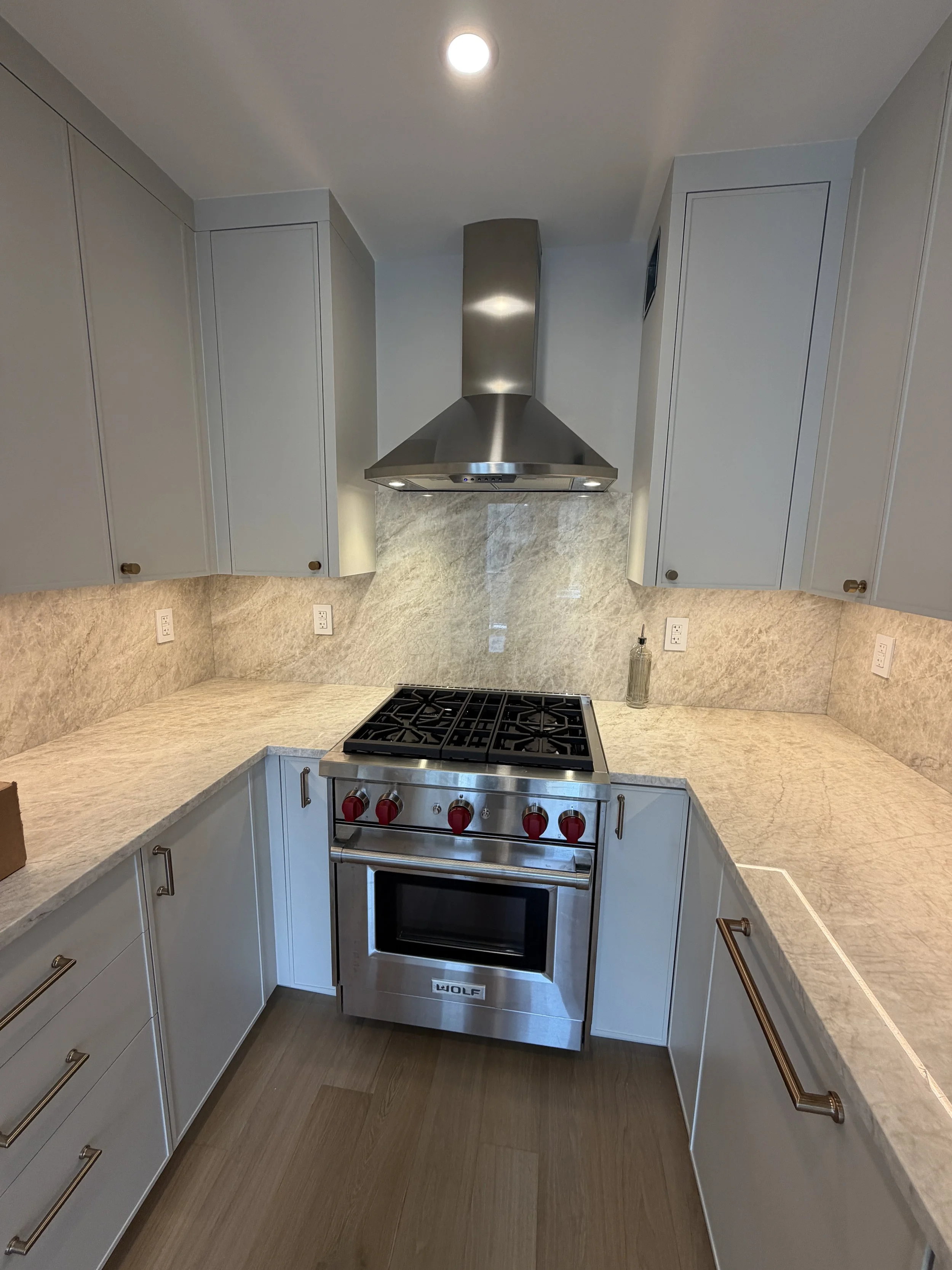 Kitchen with beige marble countertops, white cabinets, a stainless steel Wolf oven, a stainless steel range hood, and a wood floor.