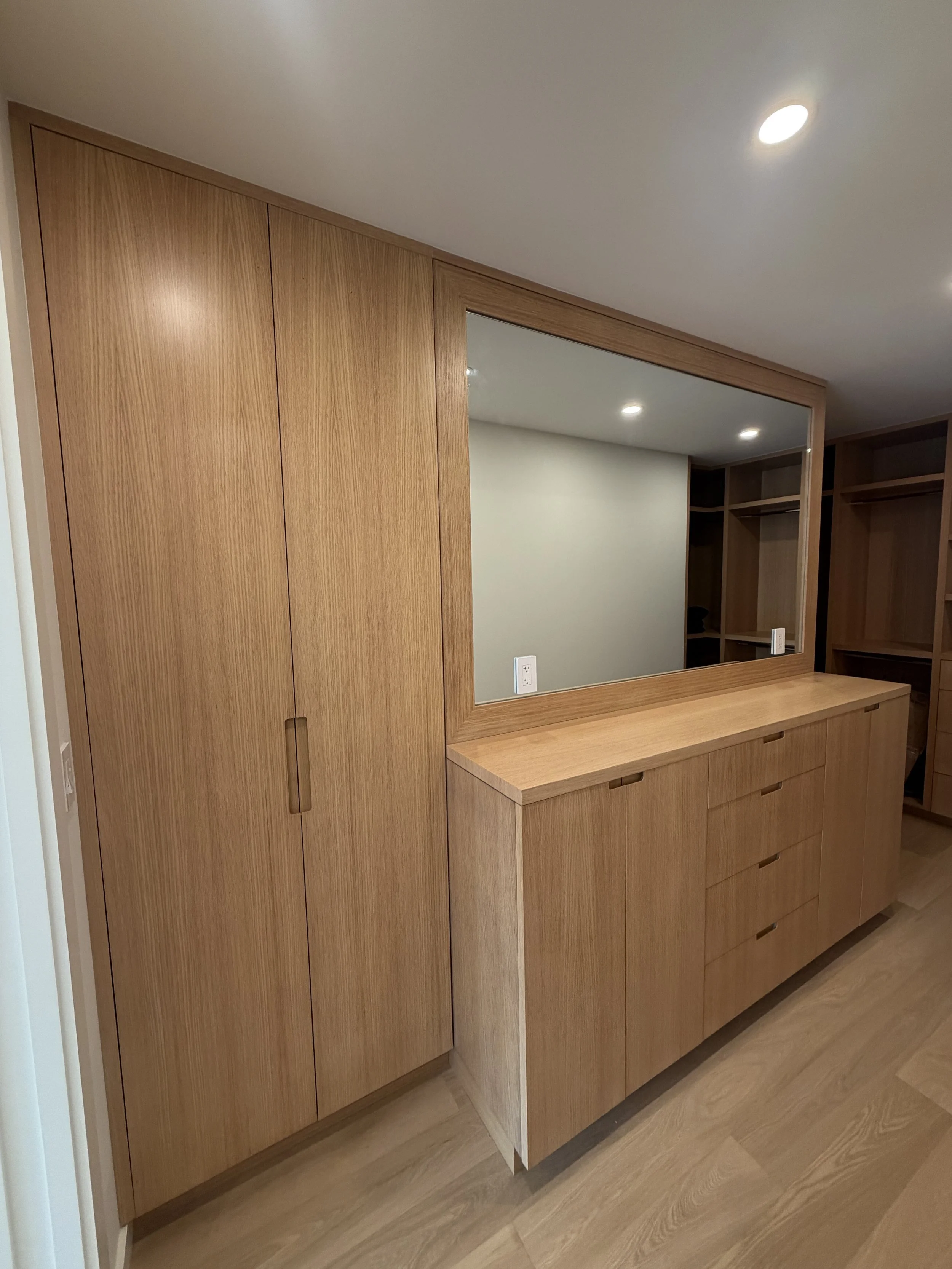A wooden cabinet with closed doors, a large mirror, and a small countertop in a room with wood flooring and ceiling lights.