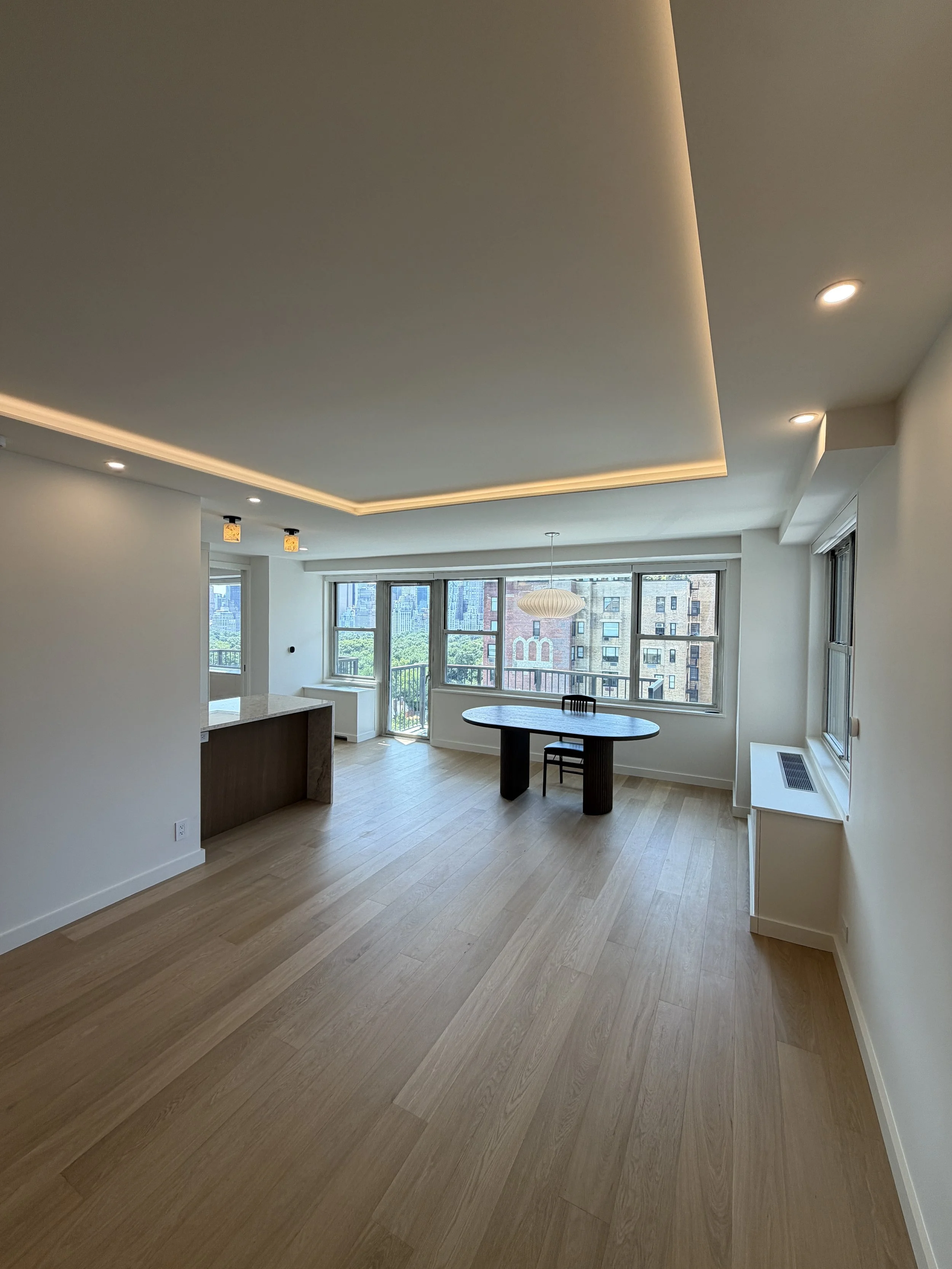 Interior of a modern apartment with hardwood floors, large windows, a dining table, and minimal decor, with city buildings visible outside.