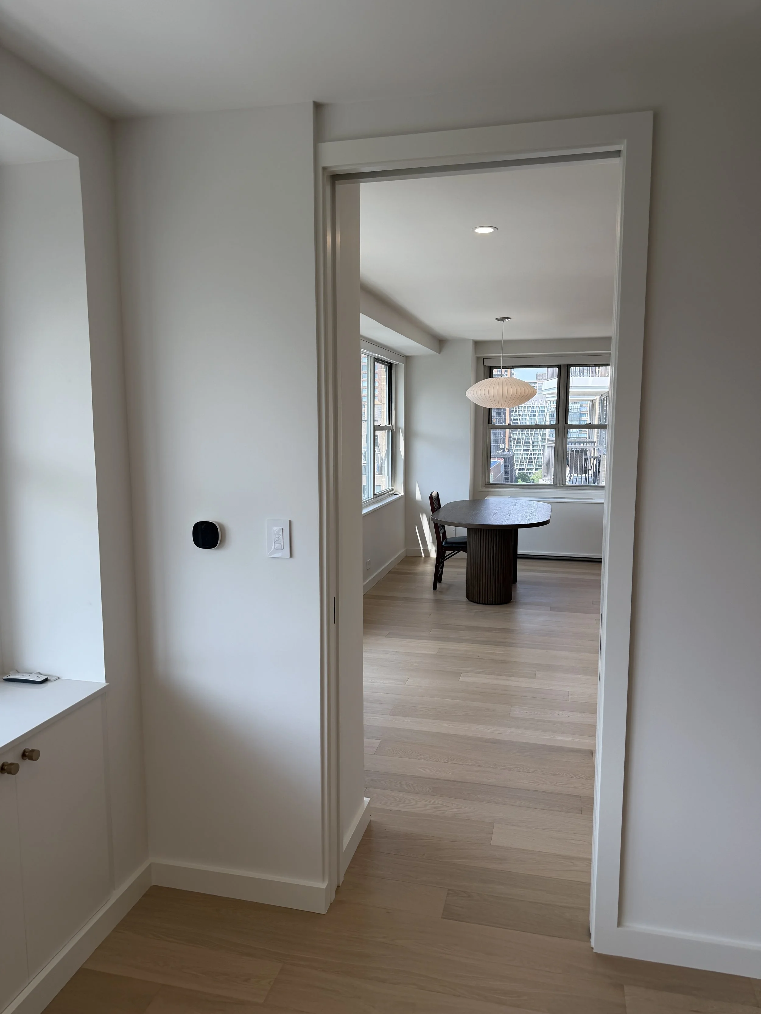 View through a doorway into a modern apartment with large windows, a small dining table, and cityscape view.