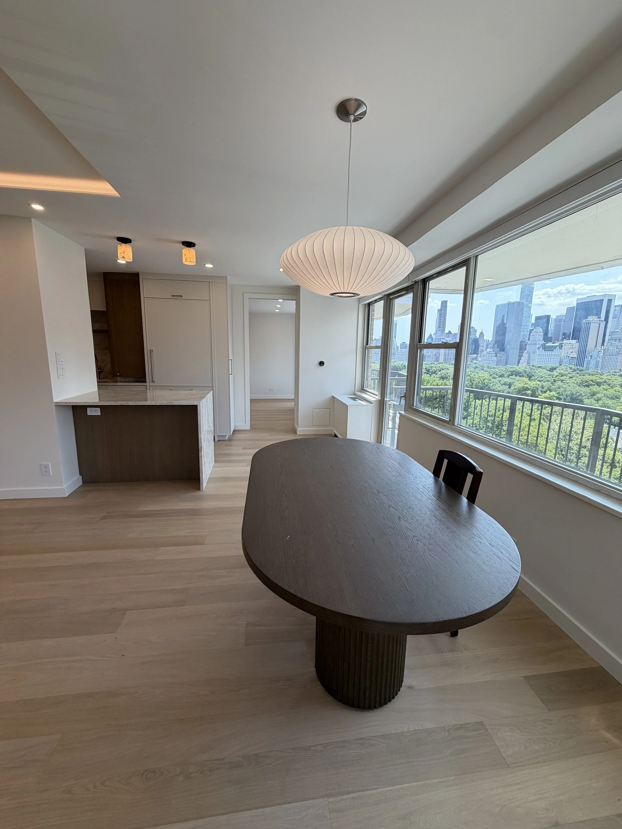 Living room with large windows showing a city skyline and green park, a hanging pendant light, and a wooden dining table with a rounded edge.