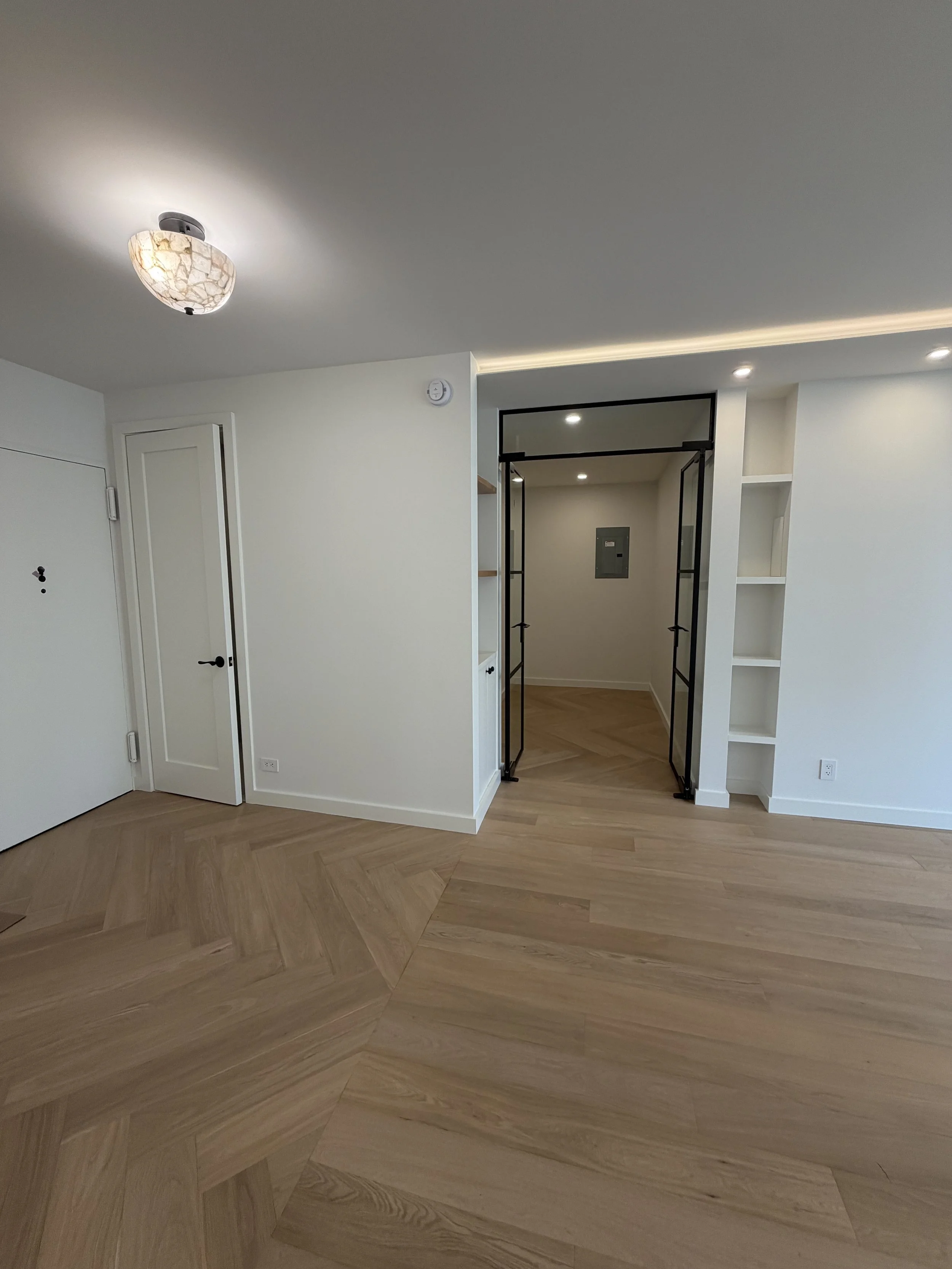 Empty modern apartment interior with white walls, wooden herringbone floor, and a doorway with black metal frame.