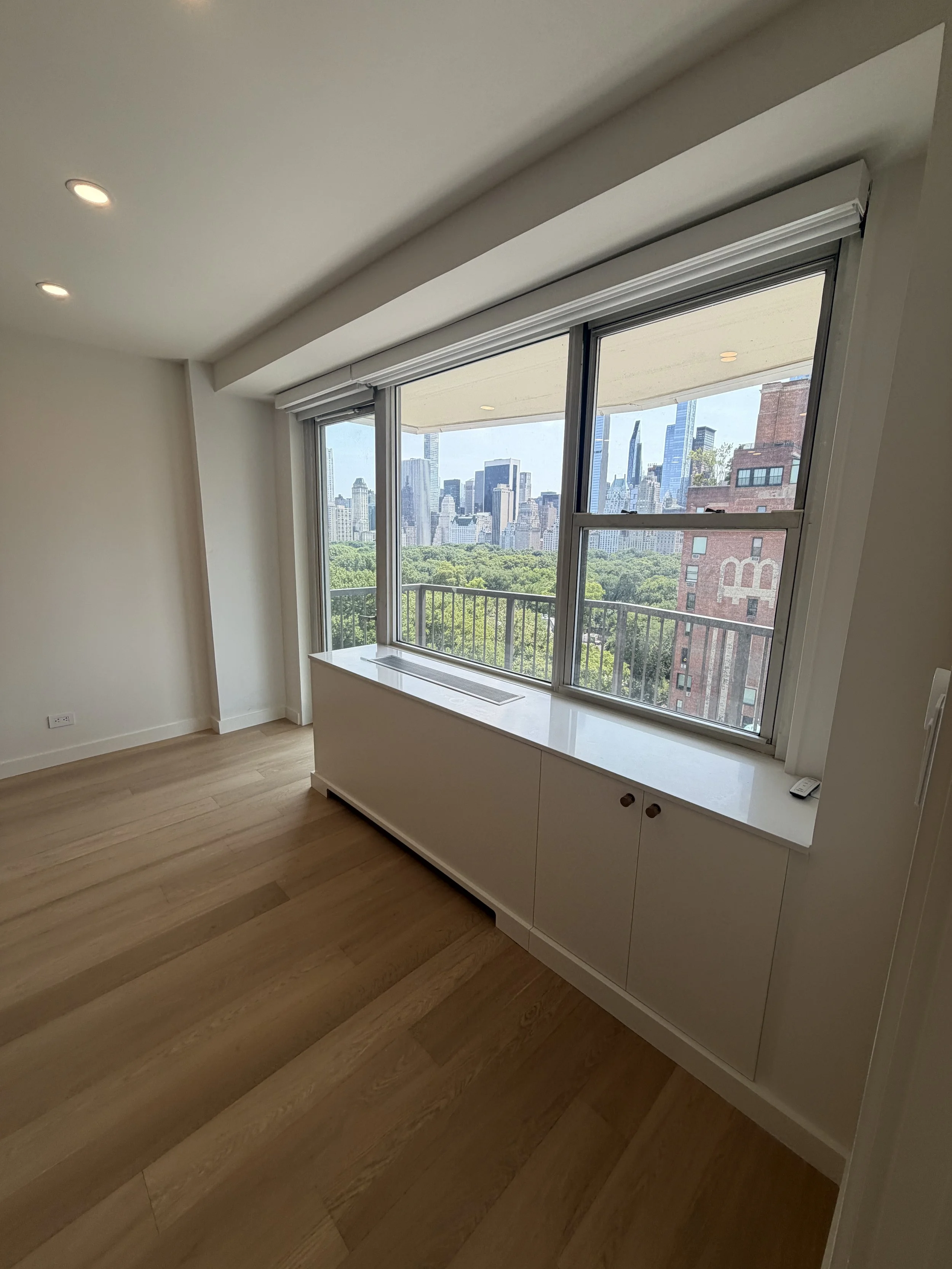 Interior of an apartment room with hardwood floors, white walls, a large window with a city skyline view, and a white cabinet below the window.