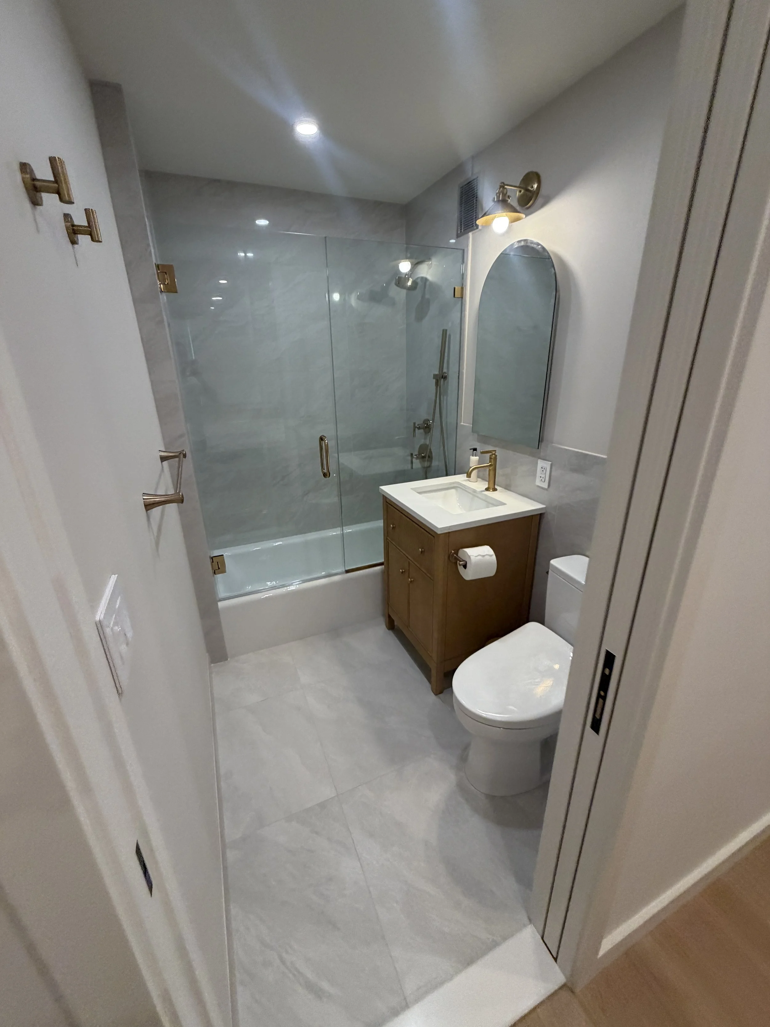 A modern bathroom with a glass-enclosed shower, a white toilet, a small wooden vanity with a sink, a mirror, and wall-mounted light fixtures.