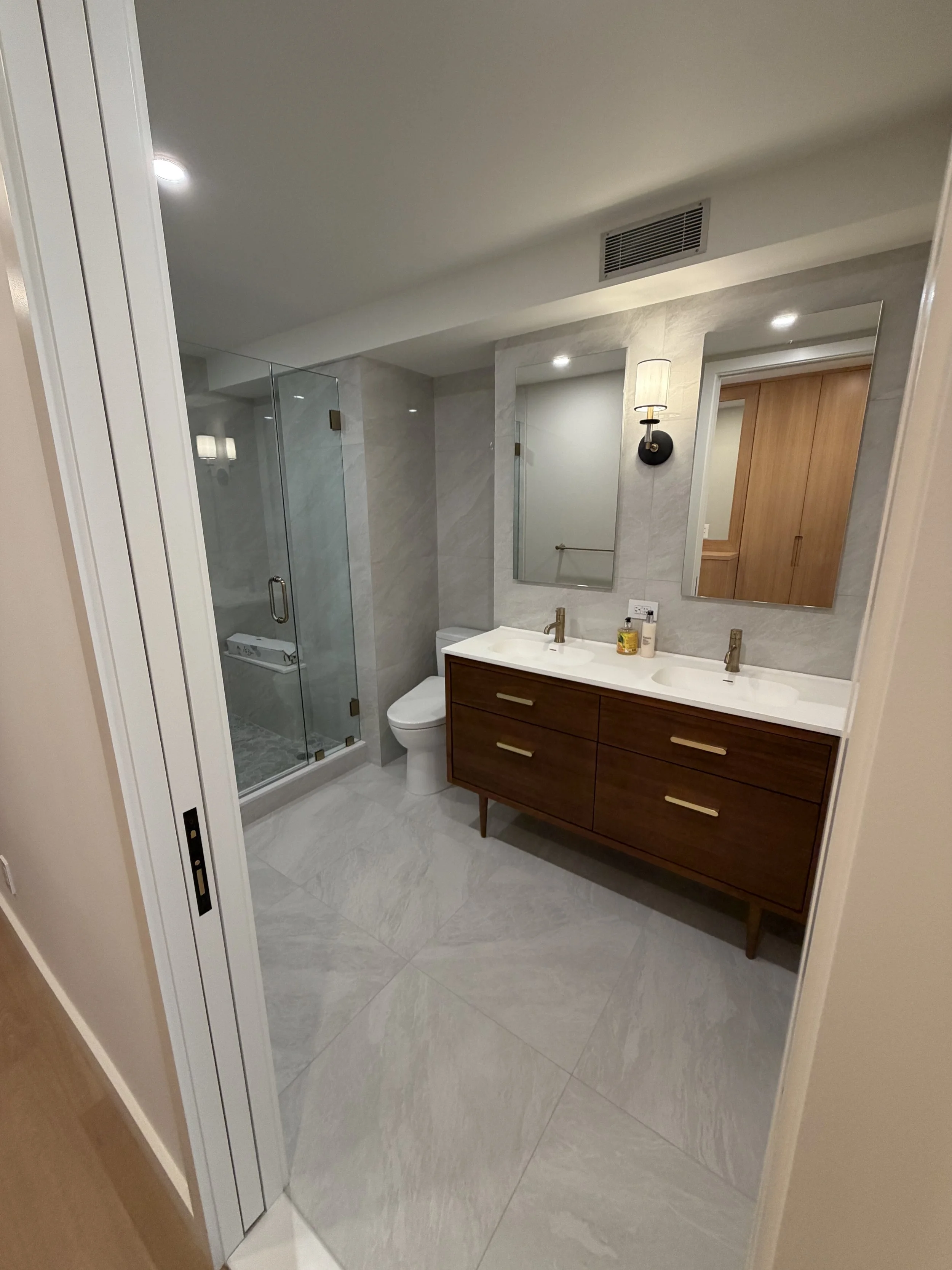 Modern bathroom with walk-in shower, double vanity with brown cabinet, two mirrors, and a wall-mounted light fixture.