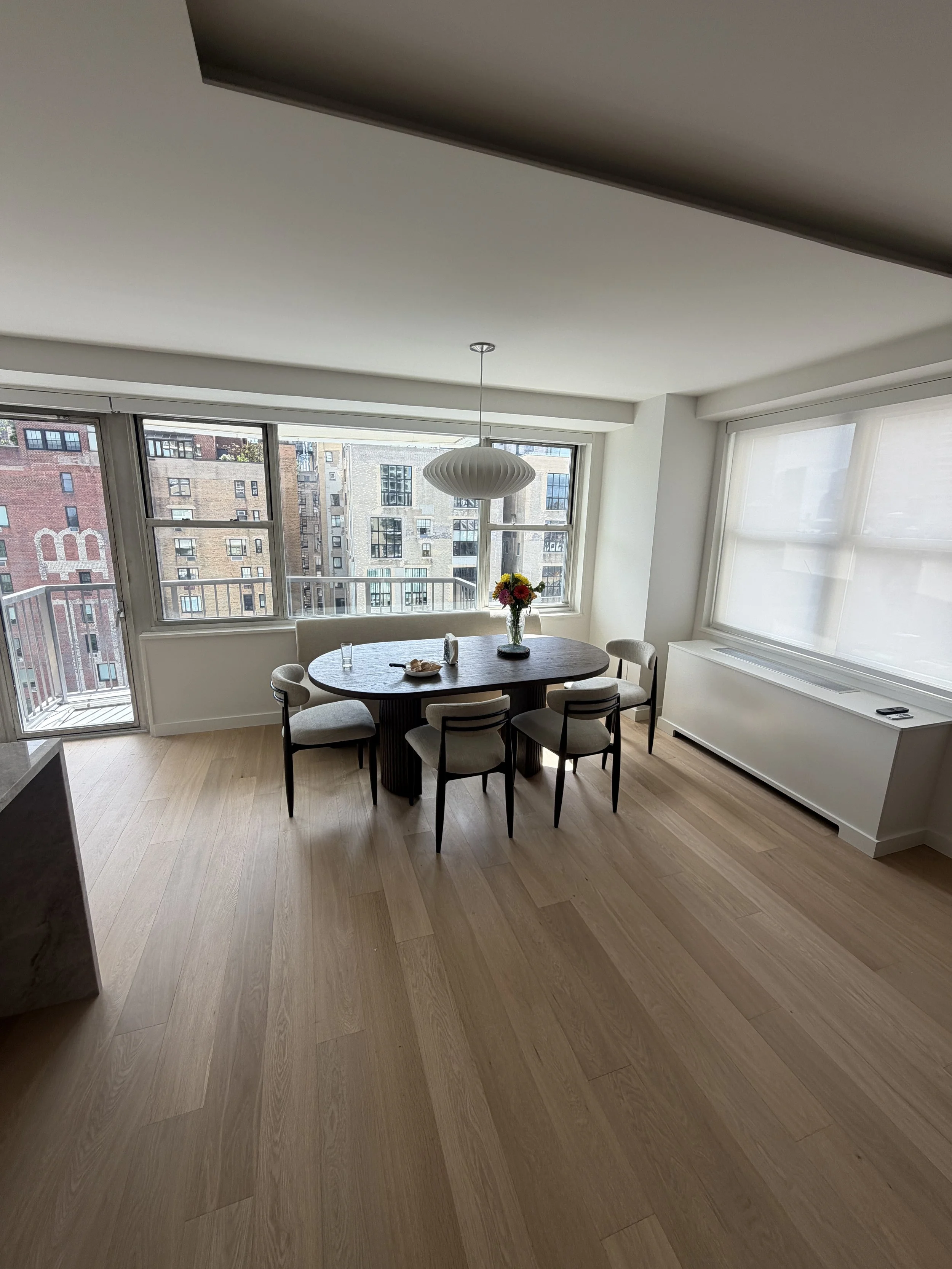Modern apartment dining area with a round table, six chairs, a vase with flowers, large windows, and a pendant light.
