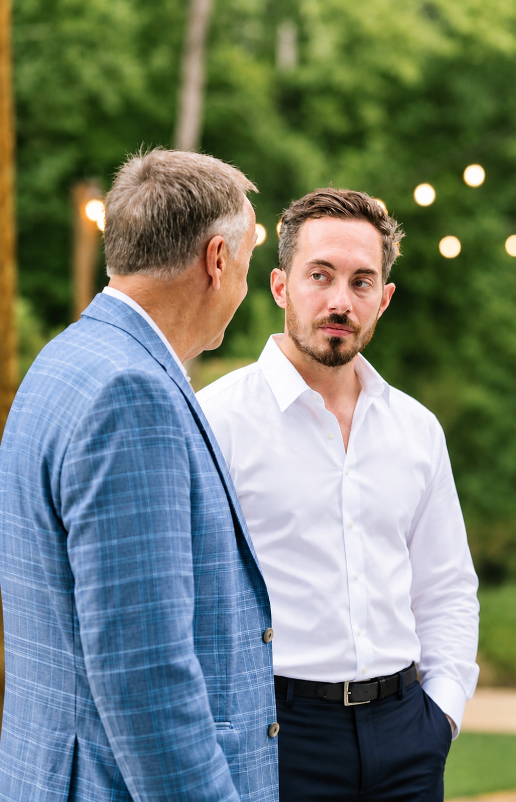 Two men are conversing outdoors at a gathering, with string lights and green trees in the background. The older man is wearing a light blue plaid suit jacket, and the younger man is in a white dress shirt with his hand in his pocket.