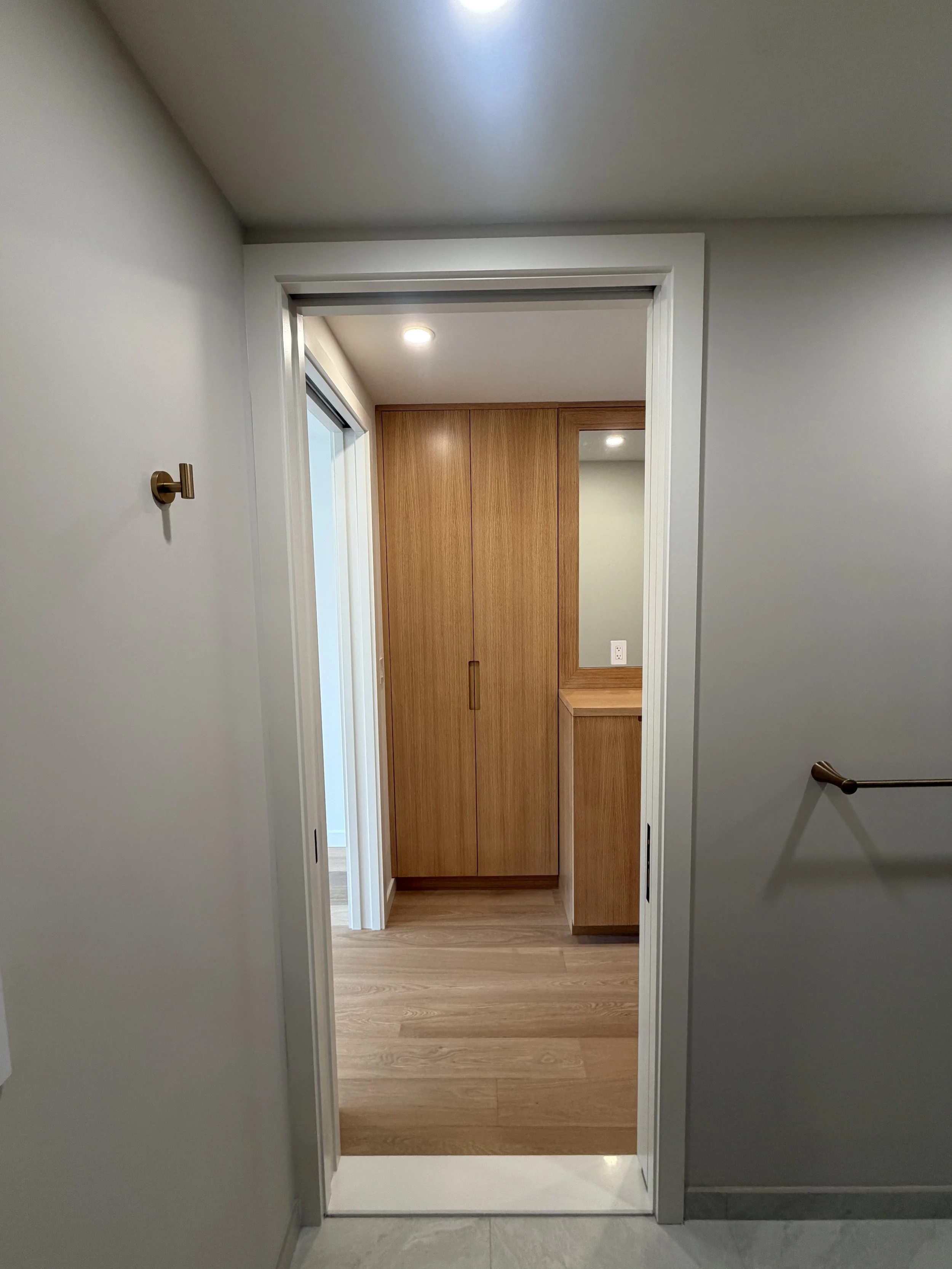 A doorway leading into a room with wooden cabinets and a wood floor. The walls are painted light gray, and there is a mirror on the right side of the room. The ceiling has a recessed light, and a brass-colored hook is installed on the left wall near 