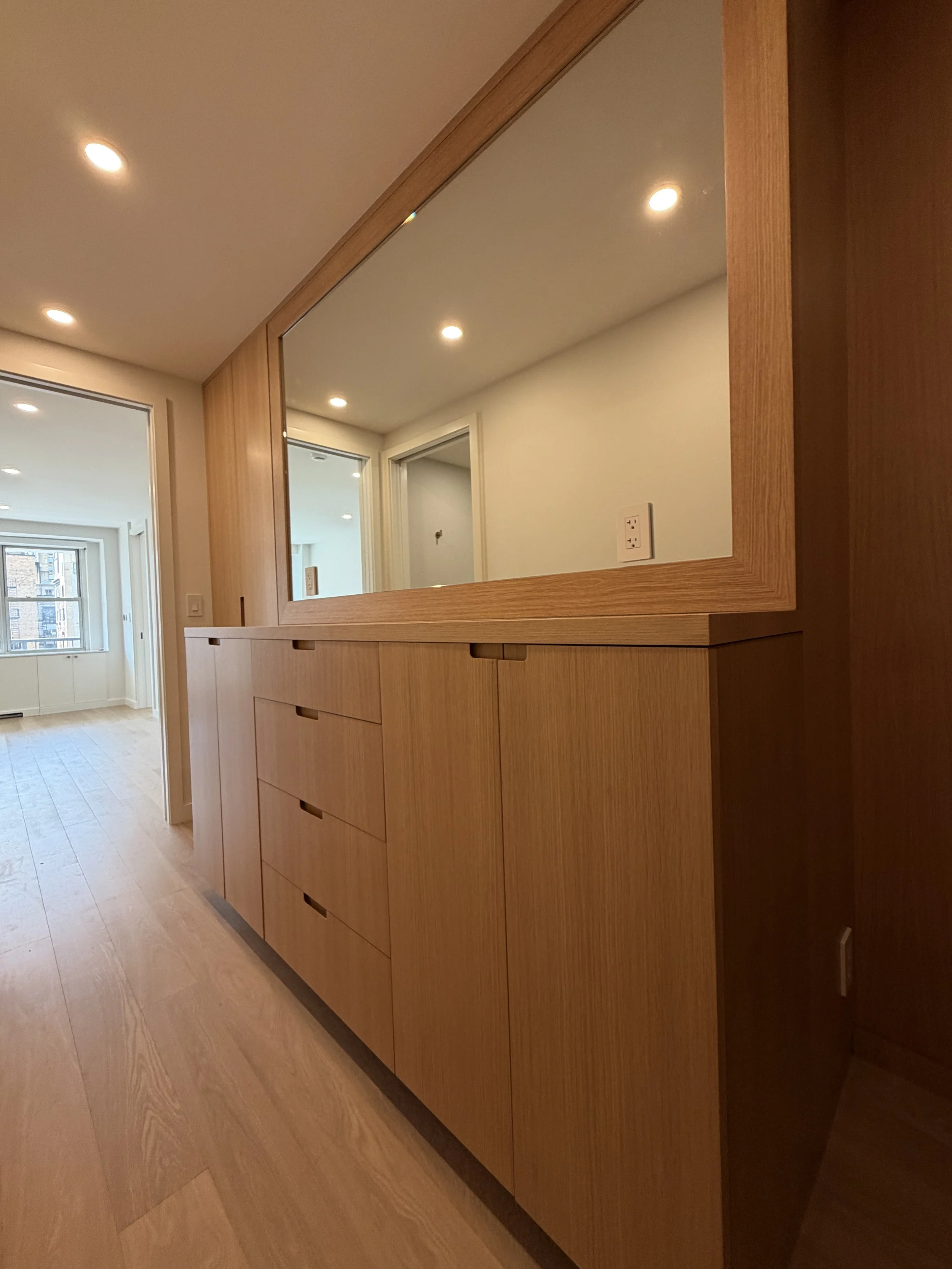 A large wooden cabinet with a mirror in the middle and drawers underneath, set against a wall with an electrical outlet. The room has light wood flooring and white walls, with a doorway leading to a bright room with windows.