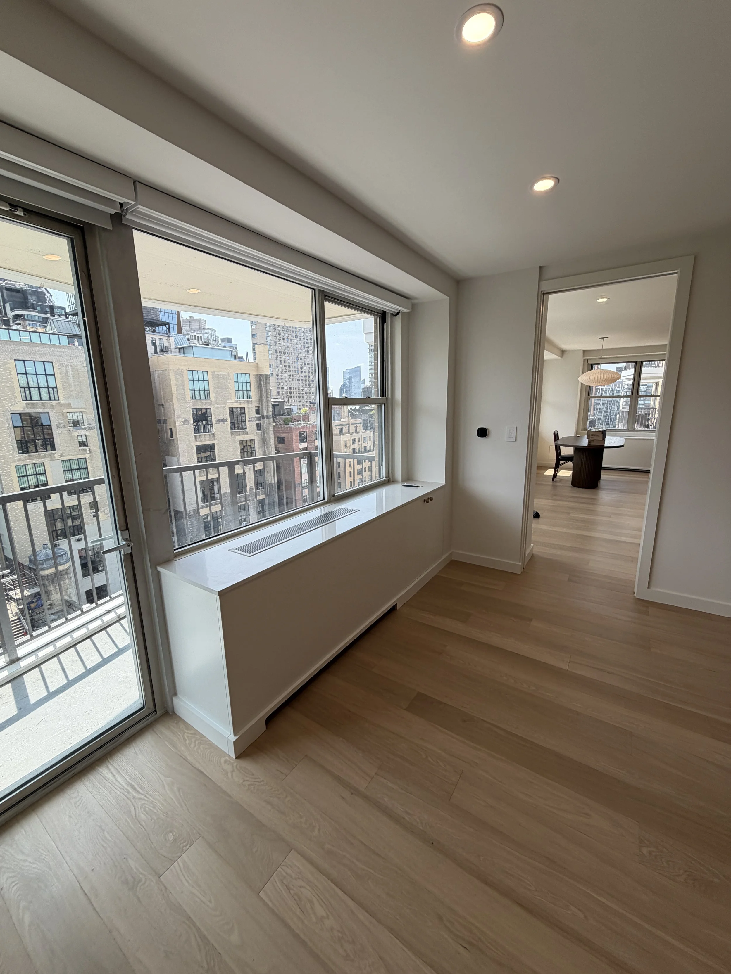 Interior view of a modern apartment with large windows showing city buildings, light wood flooring, and a round dining table with chairs in the background.