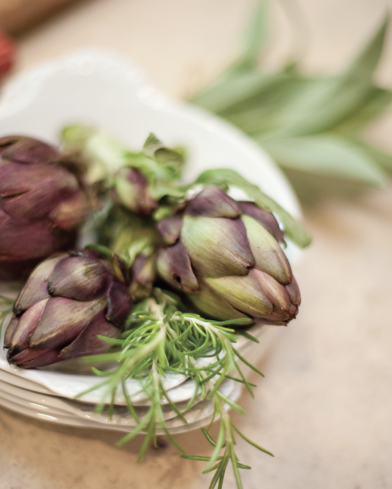 Close-up of a bunch of fresh purple artichokes with green leaves and sprigs of rosemary on a white plate.