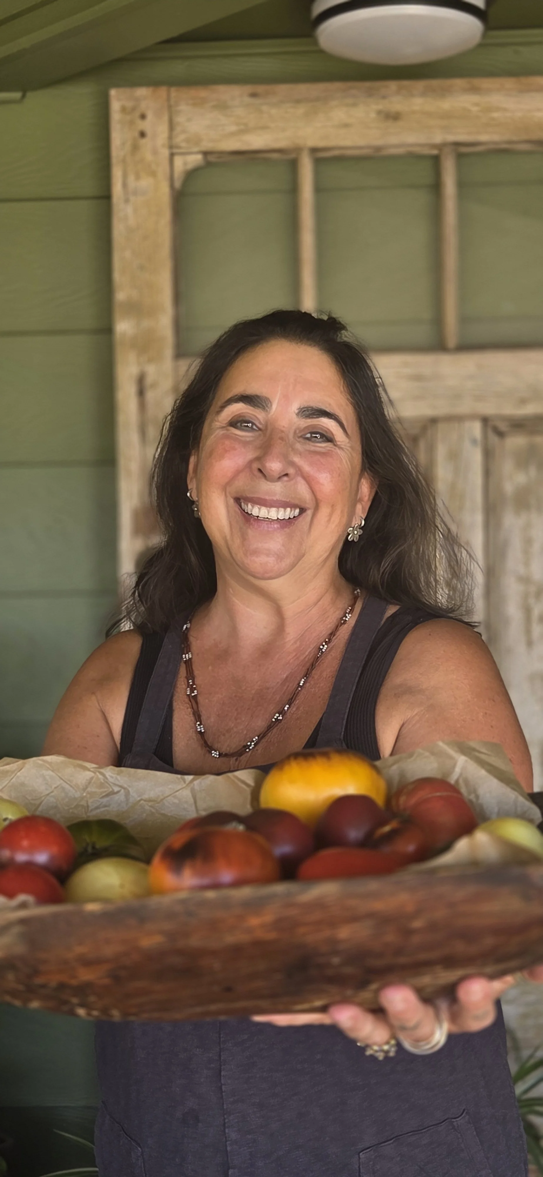 Chef Robin smiling and holding a wooden bowl filled with assorted ripe tomatoes.