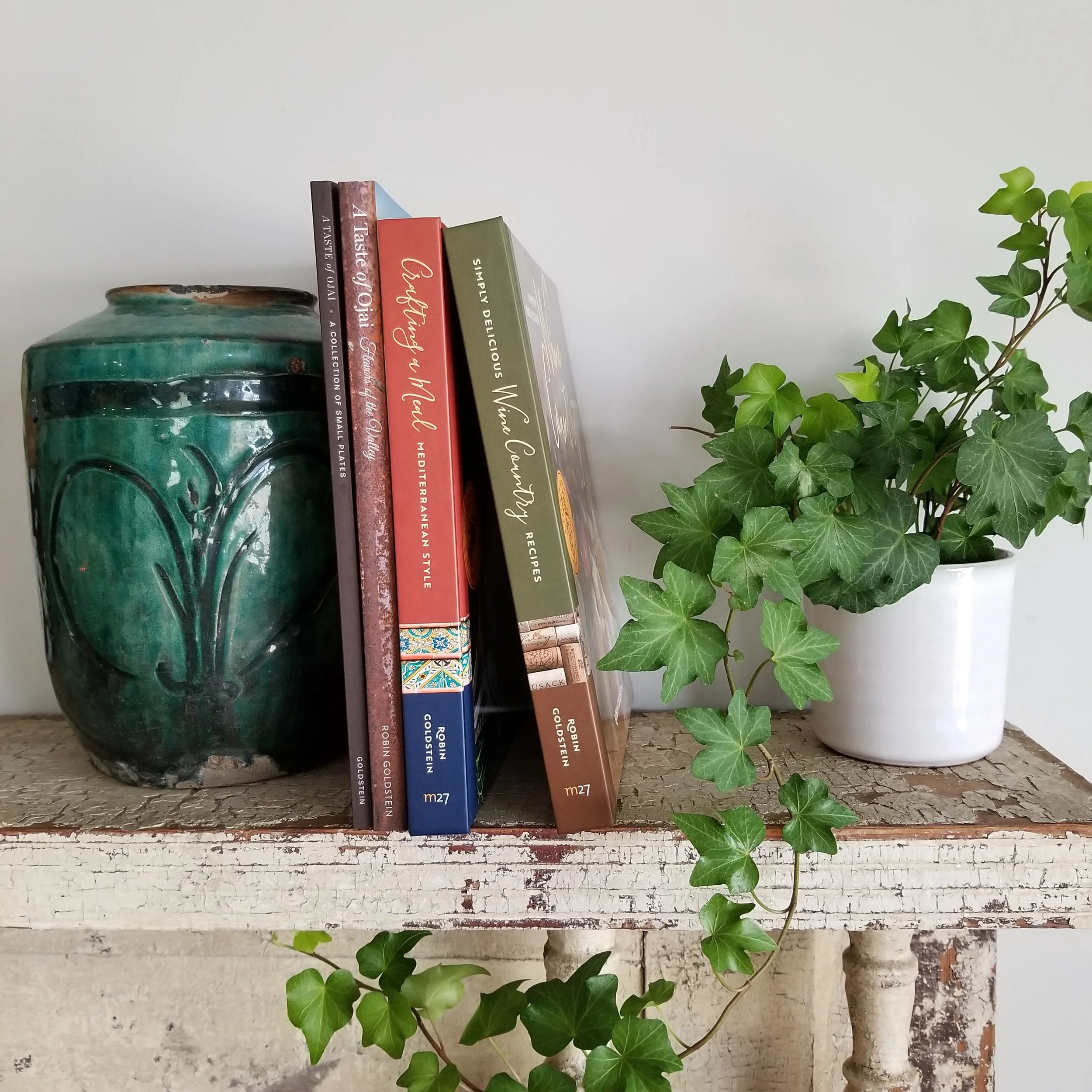 A collection of Chef Robin's cookbooks arranged vertically on a shelf, next to a potted ivy plant with green leaves, on a rustic white wooden surface.