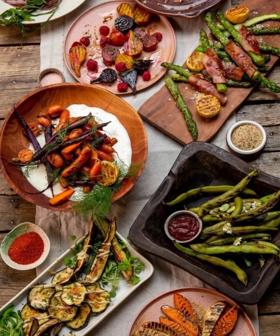 Assorted grilled vegetables and dishes on rustic wooden table, including carrots, zucchini, eggplant, asparagus, and grilled meat with dipping sauces and herbs.