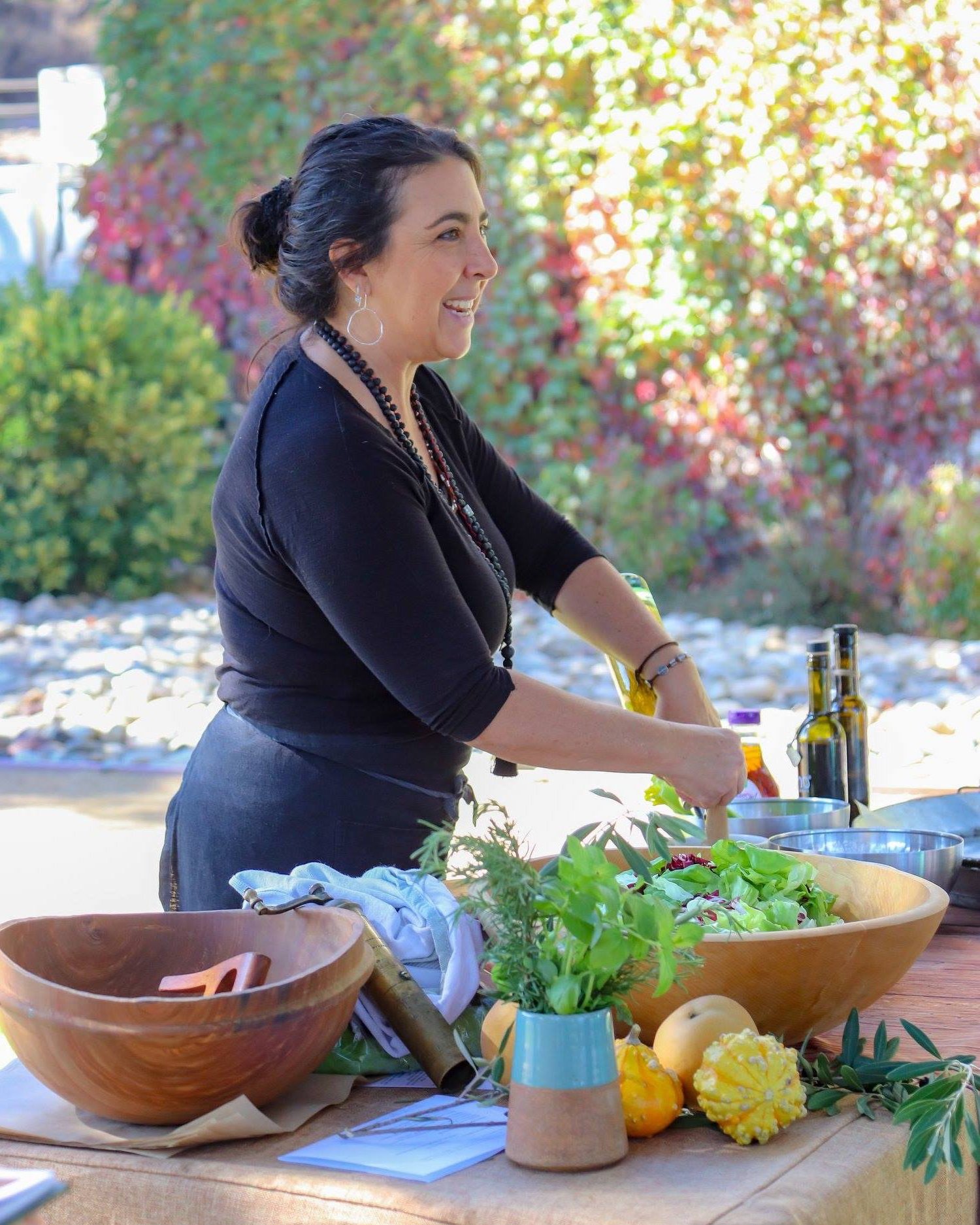 A woman with dark hair tied back, wearing black clothing and jewelry, preparing food outdoors on a table with bowls, fresh herbs, and vegetables, with colorful autumn foliage in the background.