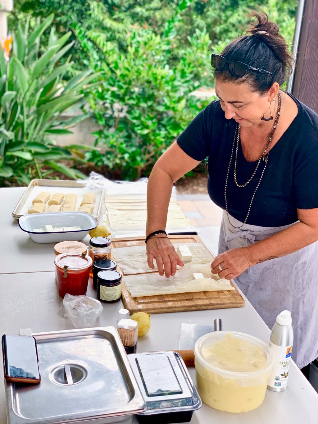 Chef Robin preparing food at outdoor cooking demo on a table with various ingredients and kitchen tools, surrounded by green plants.