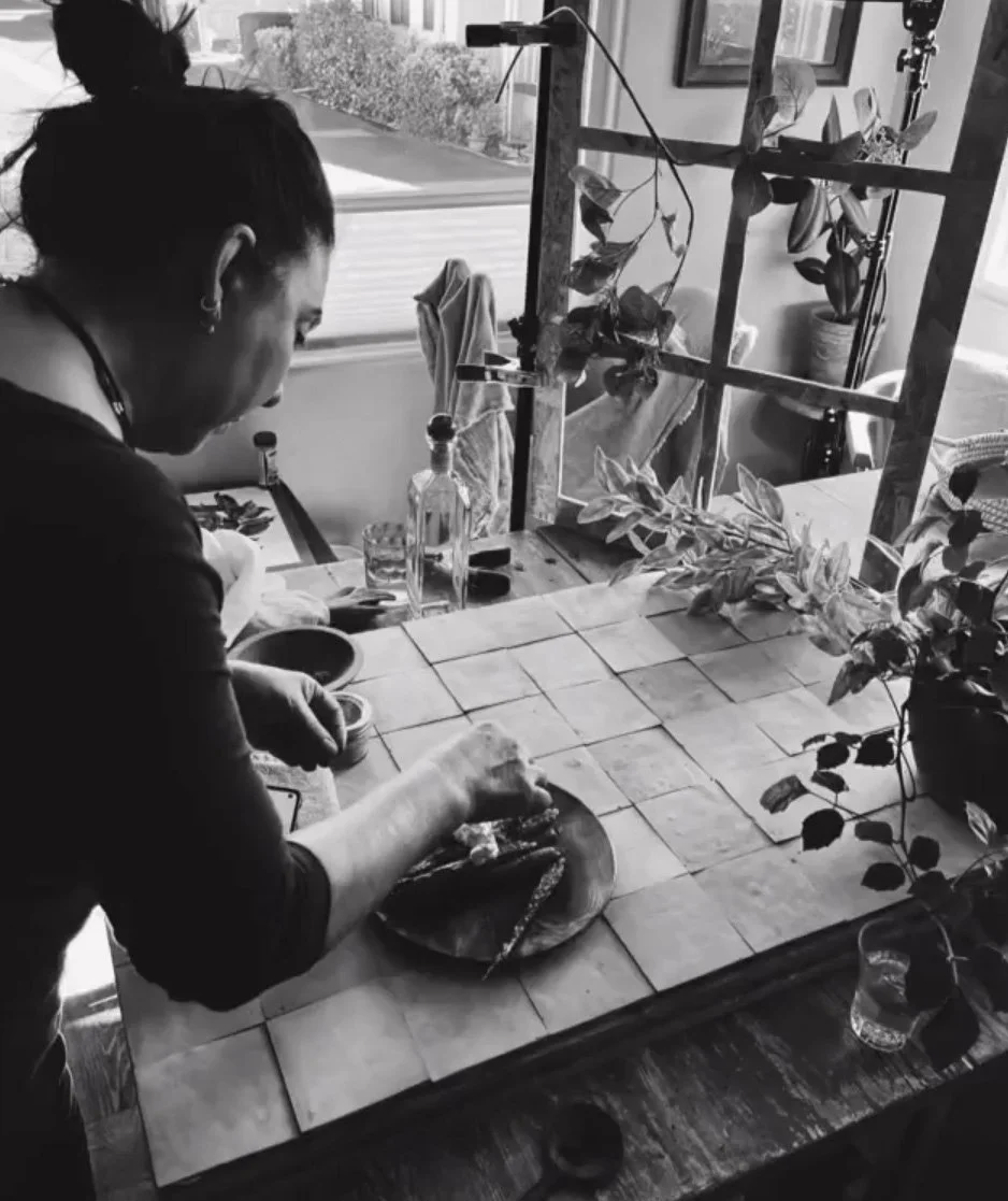 Chef Robin preparing food at a tiled kitchen counter, with plants on and near the counter, bottles and glasses nearby, outside view through the window.