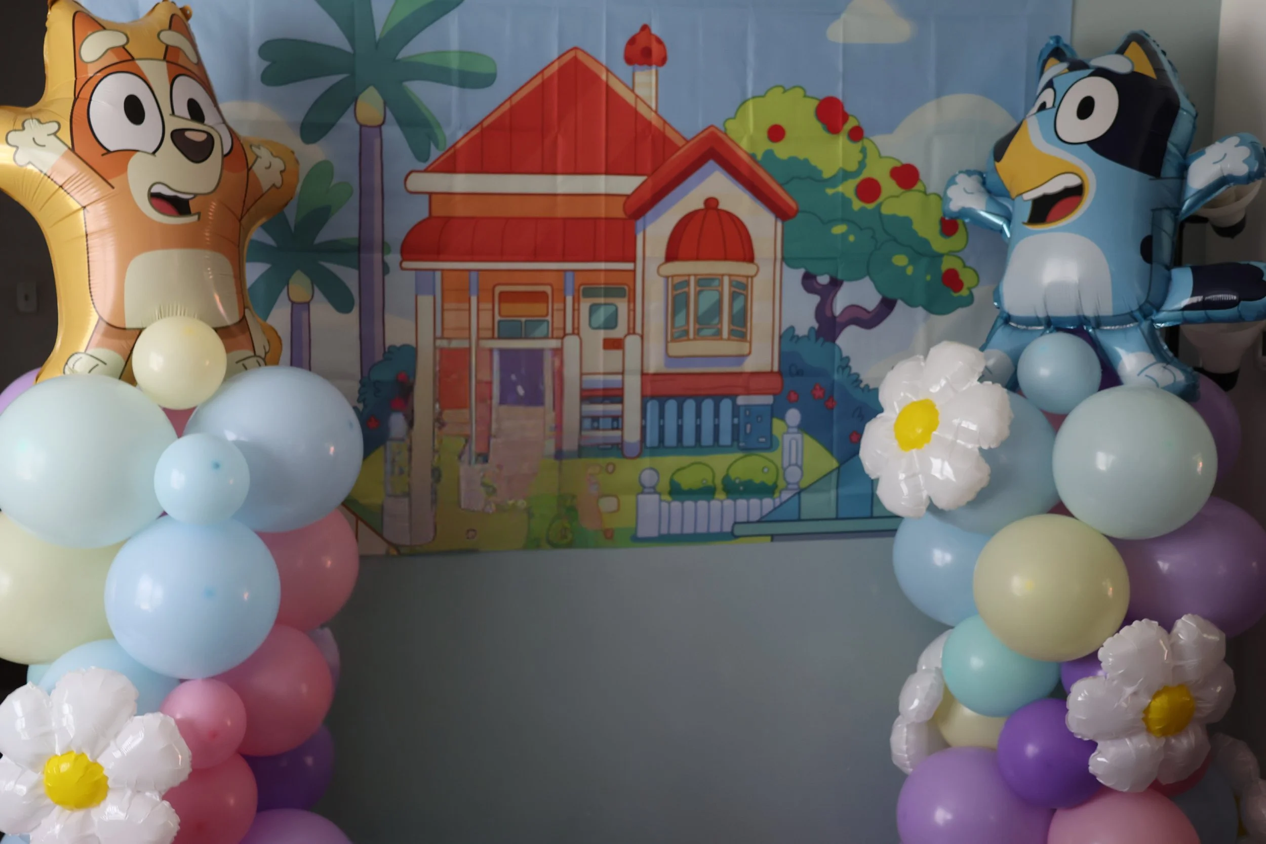Colorful balloon arrangement featuring two balloons with cartoon dog faces, one orange and one black and white, surrounding a backdrop with a house, trees, and clouds.