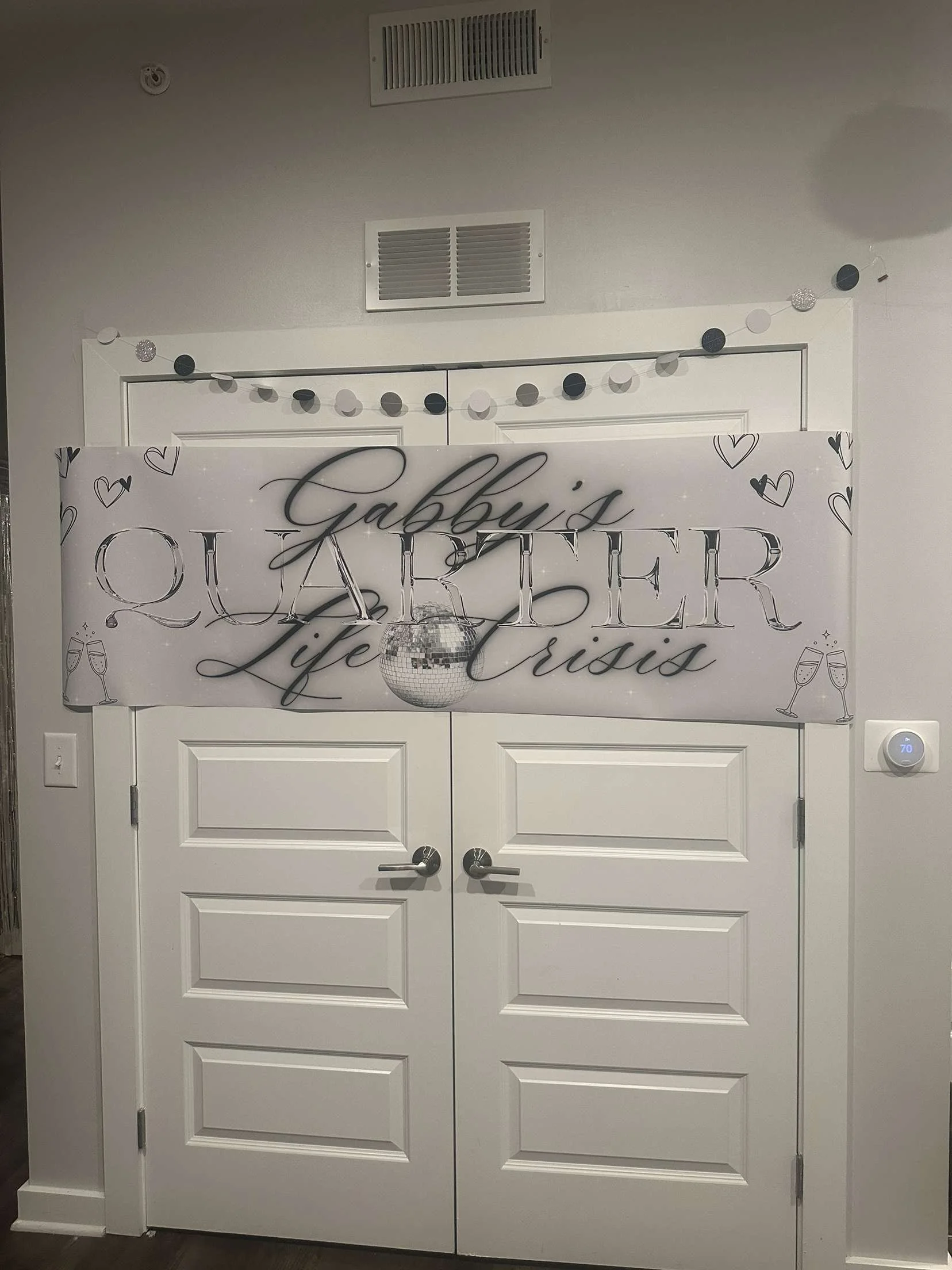 Decorated door with a sign that reads "Gabby's Quarter Life Crisis" in elegant script. A small disco ball hangs above the sign. A string of black, white, and glittery circular decorations is draped across the top of the door. 