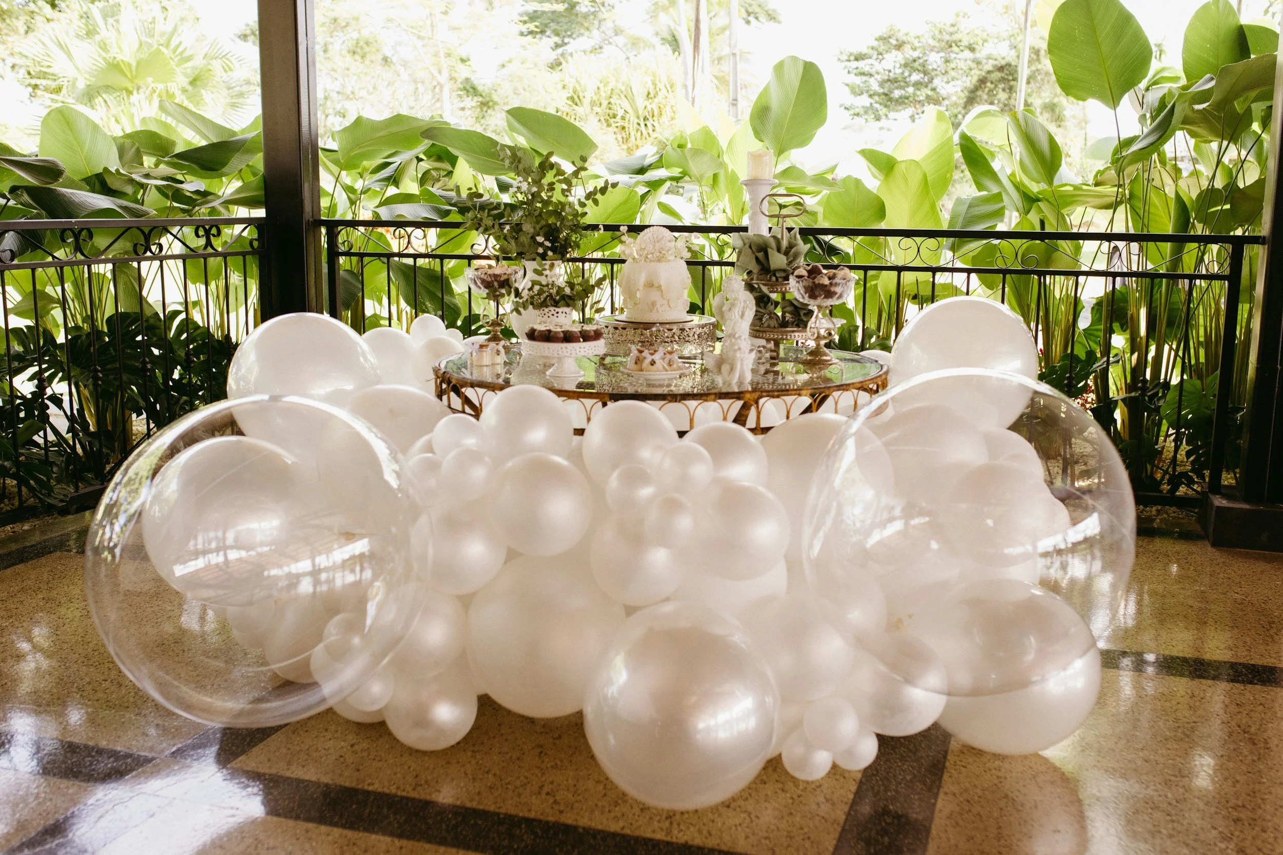 Table with white balloons in a clear container, decorated with flowers, cakes, and pastries, set outdoors with lush green plants in the background.