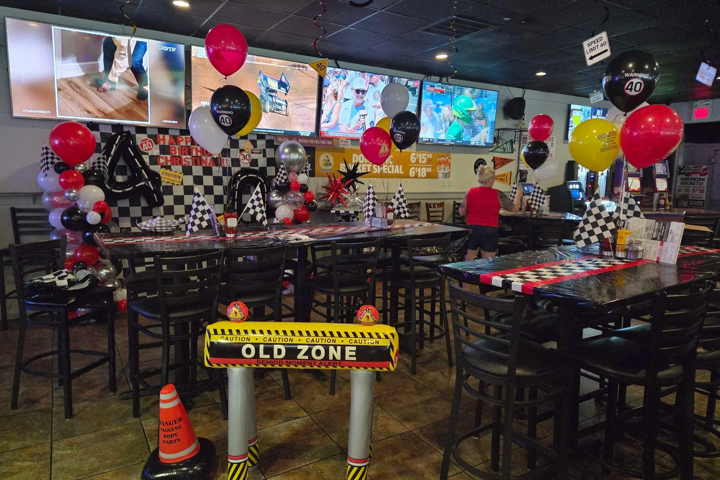 Empty restaurant decorated with racing-themed balloons and checkered flags for a birthday celebration, with television screens showing races and a woman at a table.