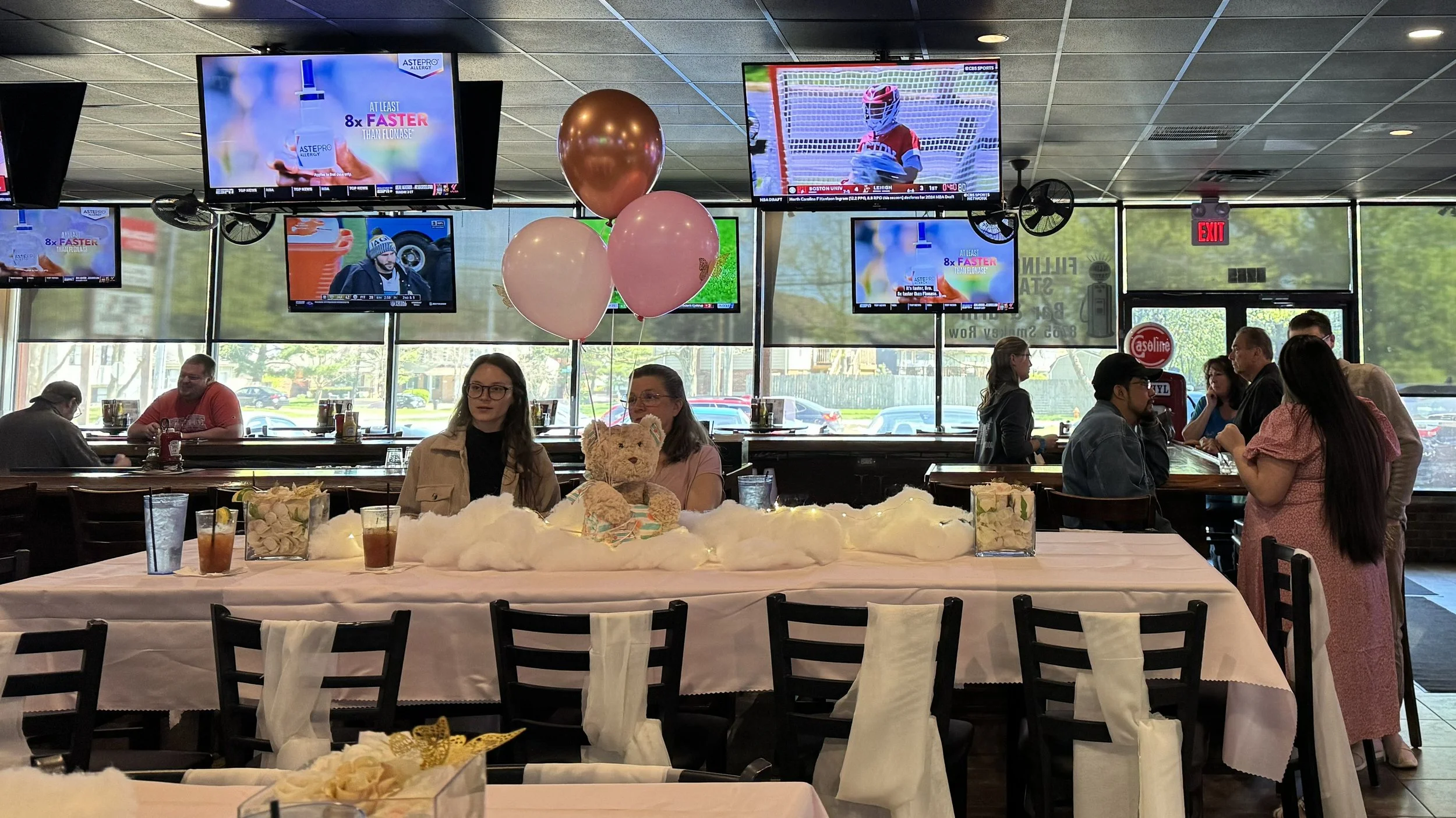 A group of people in a restaurant celebrating a baby shower with a table decorated with pink balloons, teddy bear, and a cake. Multiple television screens display sports and advertisements. Sunlight streams through large windows.