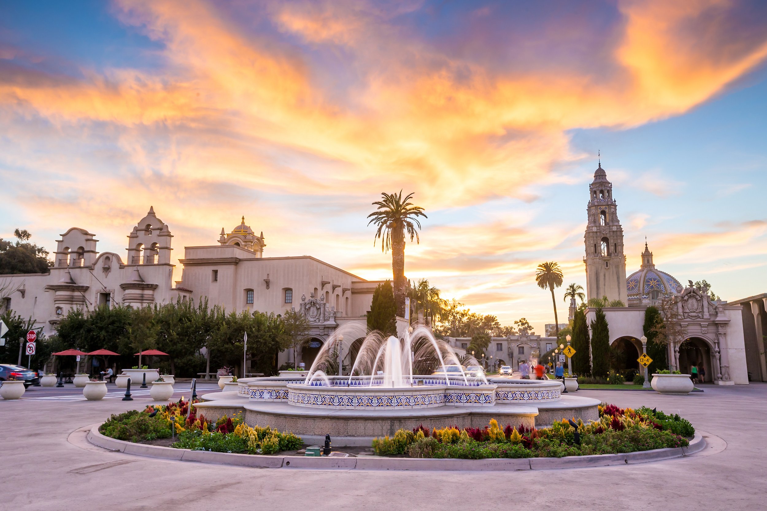 Balboa Park's large decorative fountain with water jets in the foreground surrounded by a circular flower bed, with historic Spanish-style buildings, tall palm trees, and a colorful San Diego sunset sky with orange and pink clouds in the background.