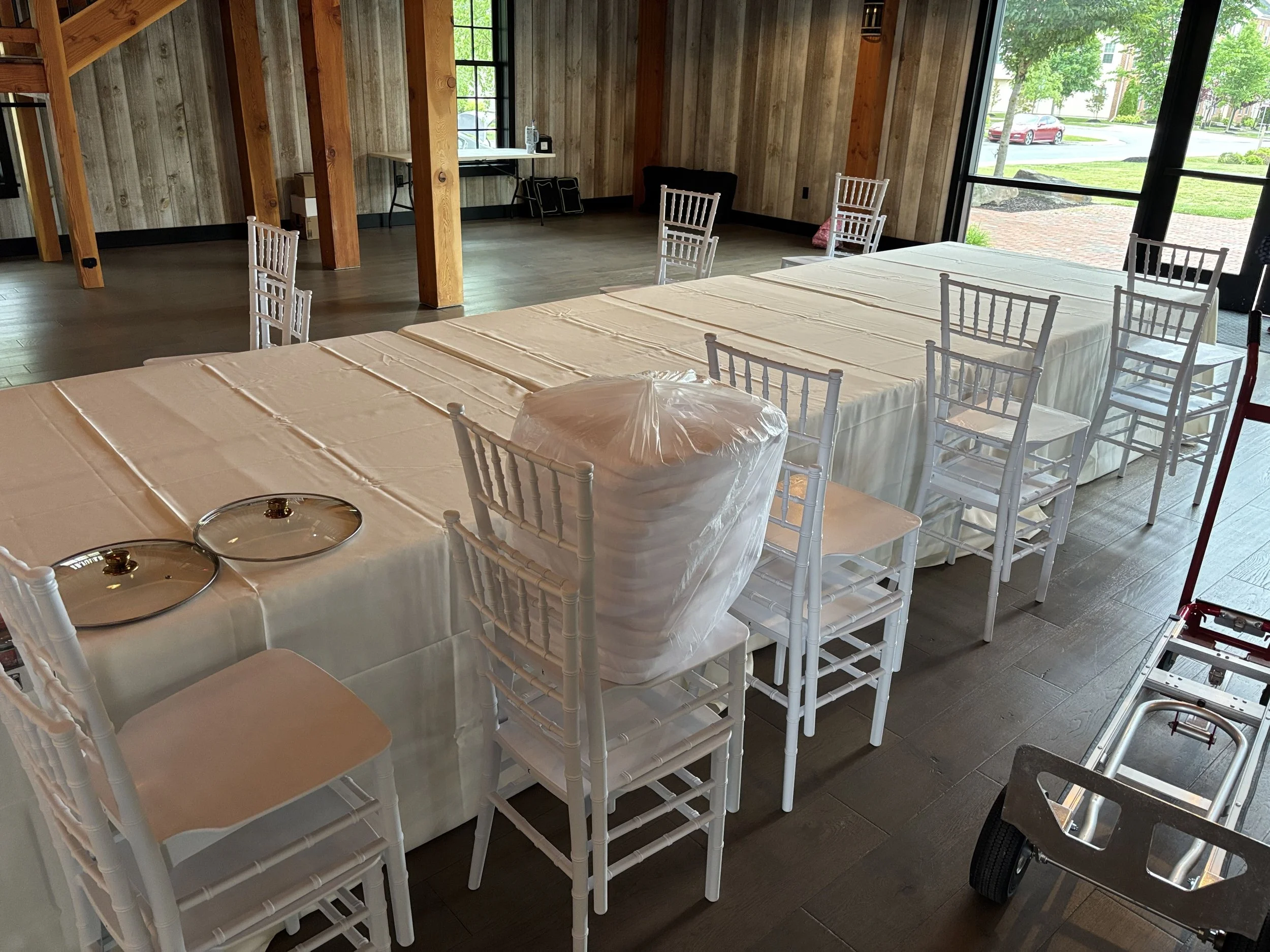 Room with long table covered in a white tablecloth, surrounded by white chairs, bright windows showing a view of green trees and a red car outside.