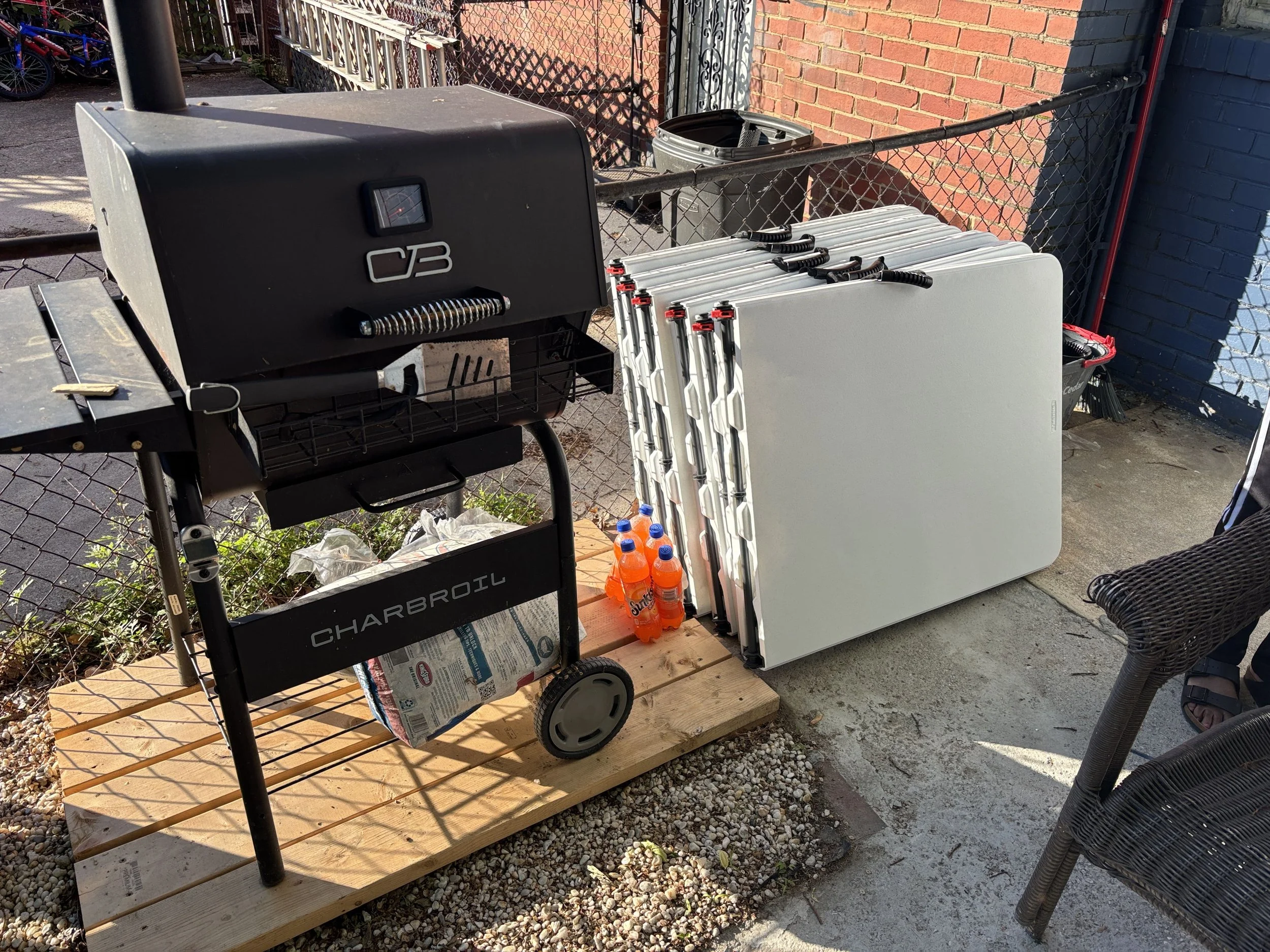 Outdoor scene showing a barbecue grill, a cart with four gas cans, a stack of white folding chairs, and a black trash can, all placed on a wooden platform next to a chain-link fence and brick wall.