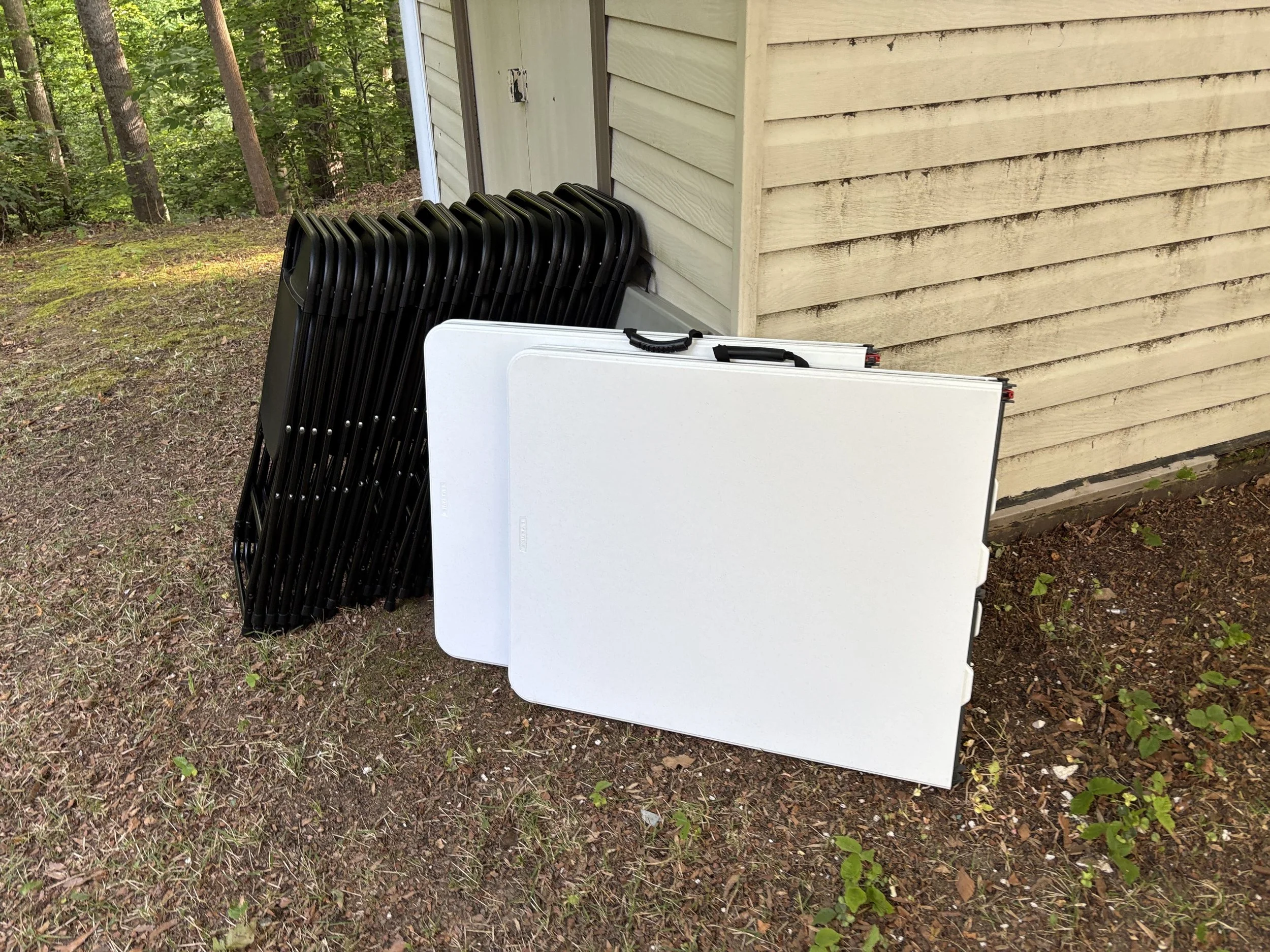 Black folding camping chairs leaning against a light-colored wooden wall outside near trees and shrubs on a dirt ground.