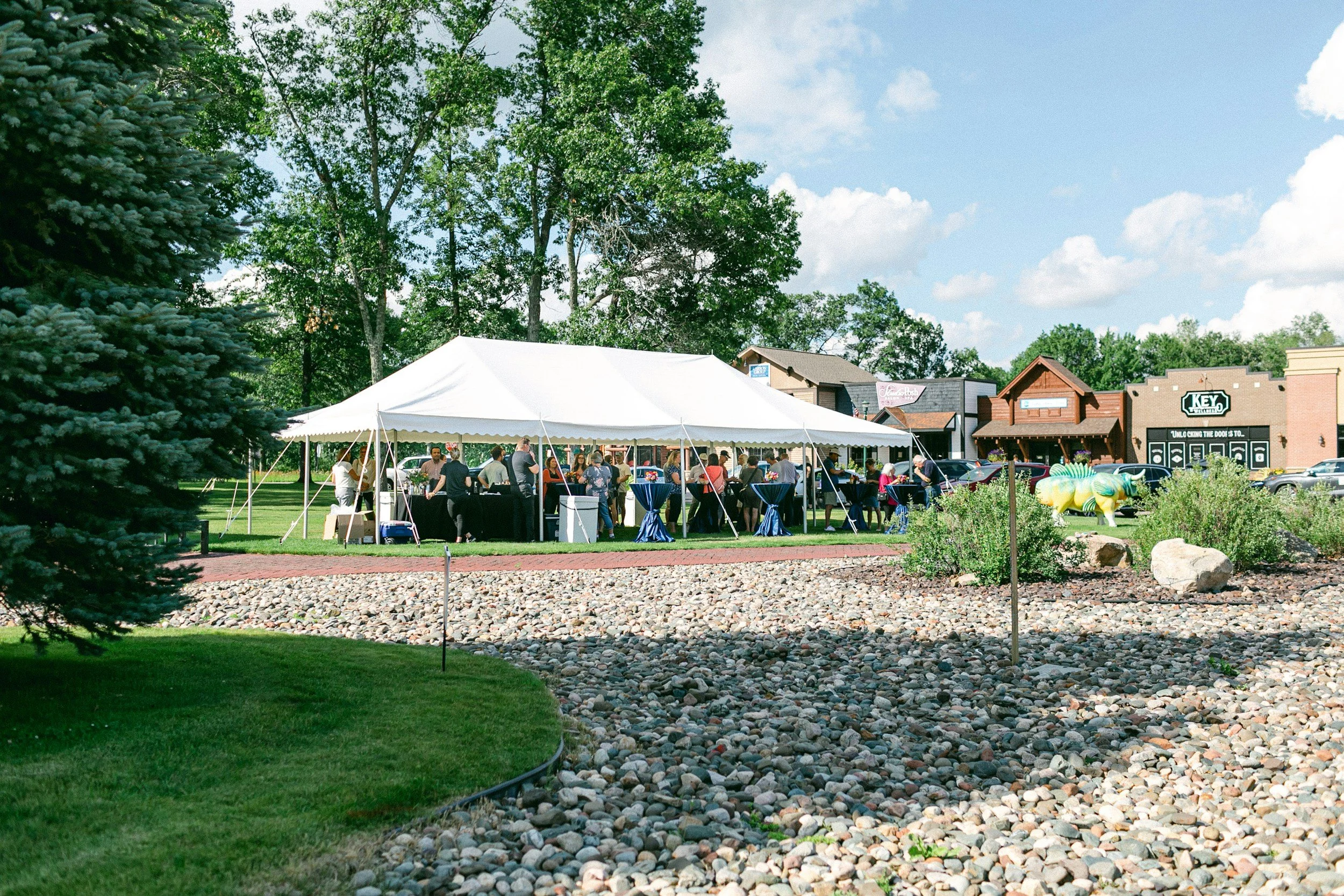 A large white event tent set up on a grassy area with people gathering underneath, surrounded by trees and small buildings in the background, with a colorful sculpture of a dinosaur and a sign on the building detecting a family-friendly outdoor event.