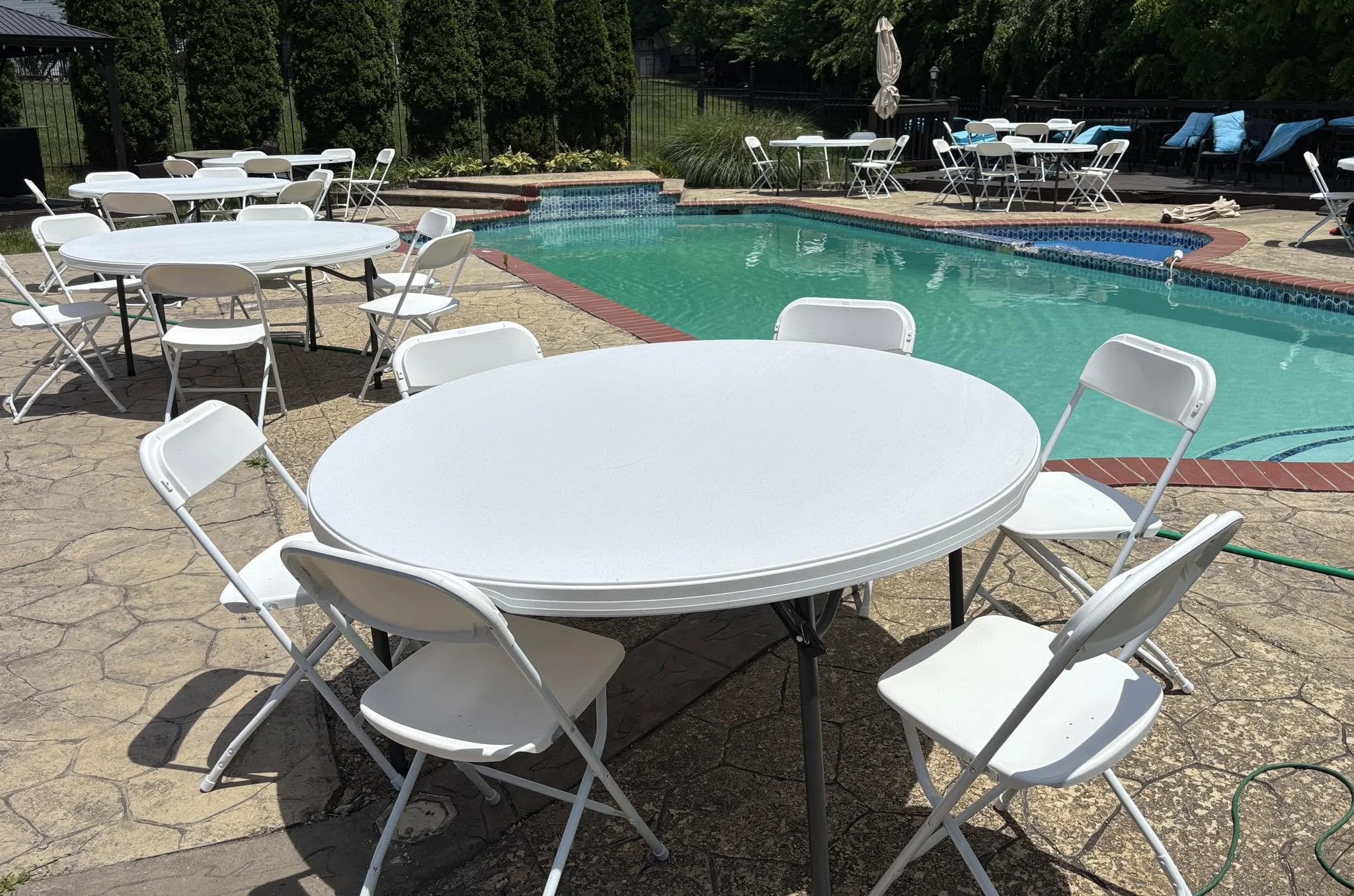 Empty white foldable chairs and round tables set up around a swimming pool on a patio, with trees and lounge chairs in the background.