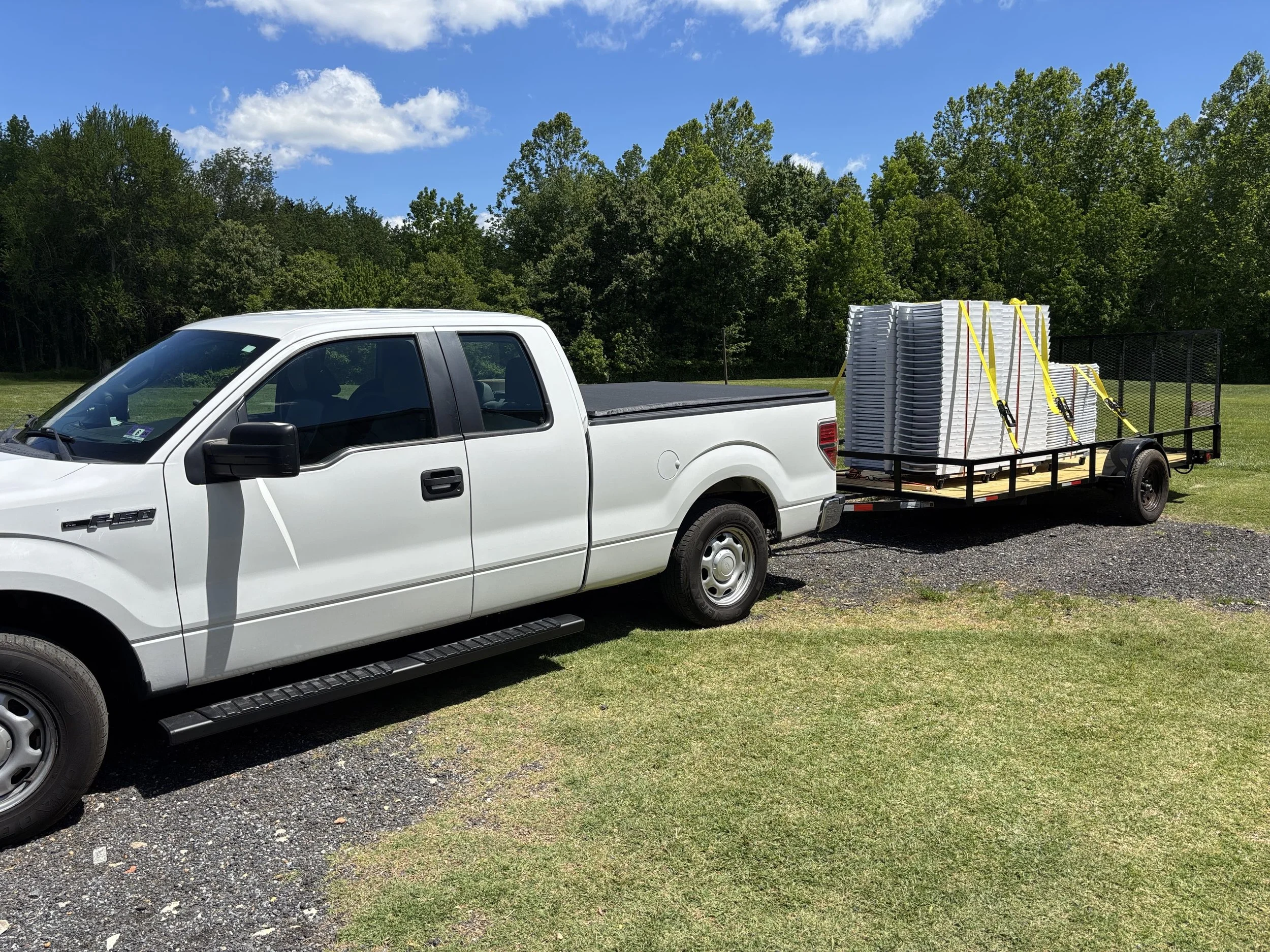 A white pickup truck with a trailer attached, carrying stacked white plastic chairs secured with yellow straps, parked on a grassy area with trees and a bright blue sky in the background.