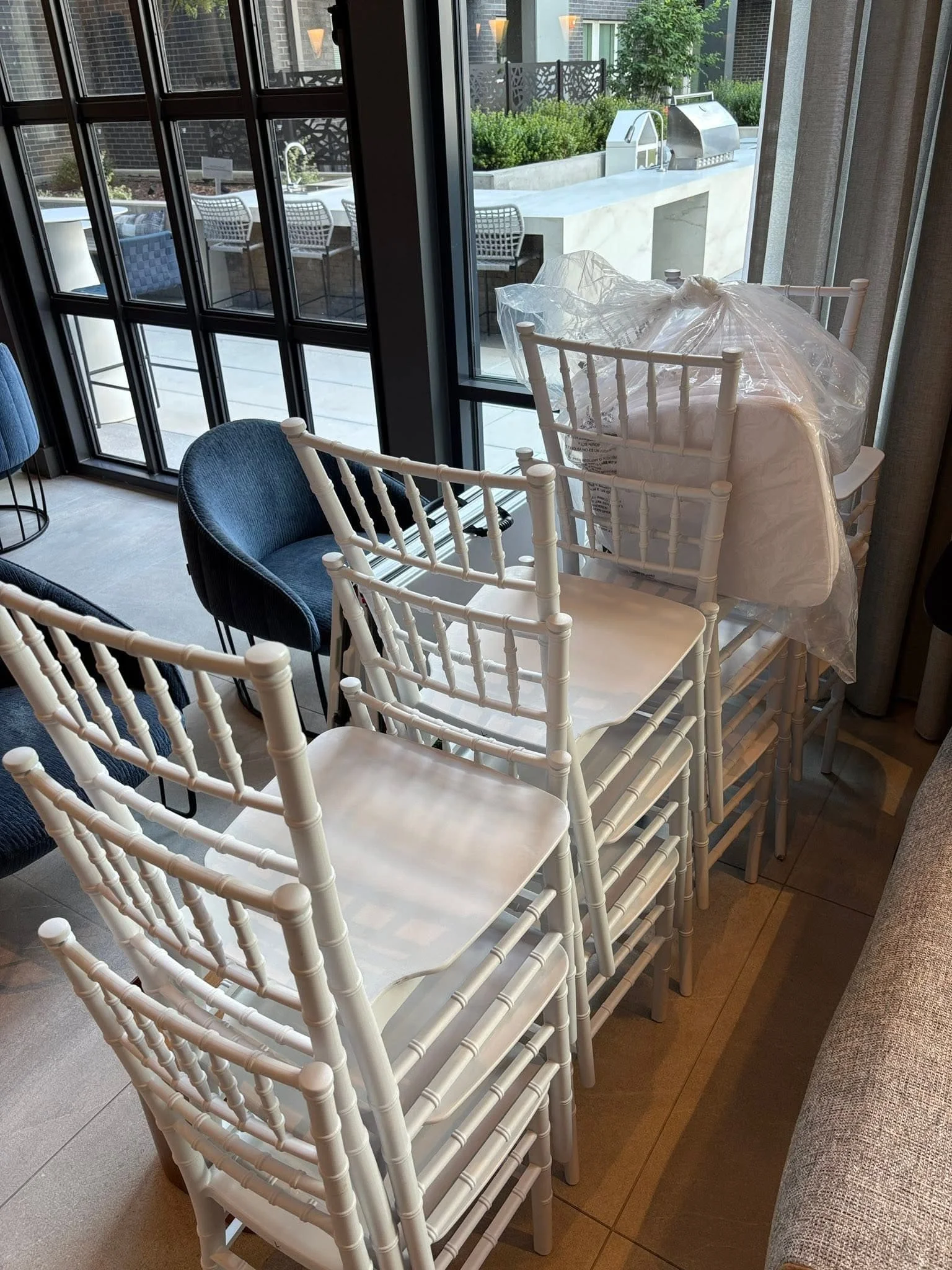 A dining area with white chairs stacked around a table covered with a clear plastic sheet, located near a large window overlooking an outdoor patio with chairs, tables, and greenery.