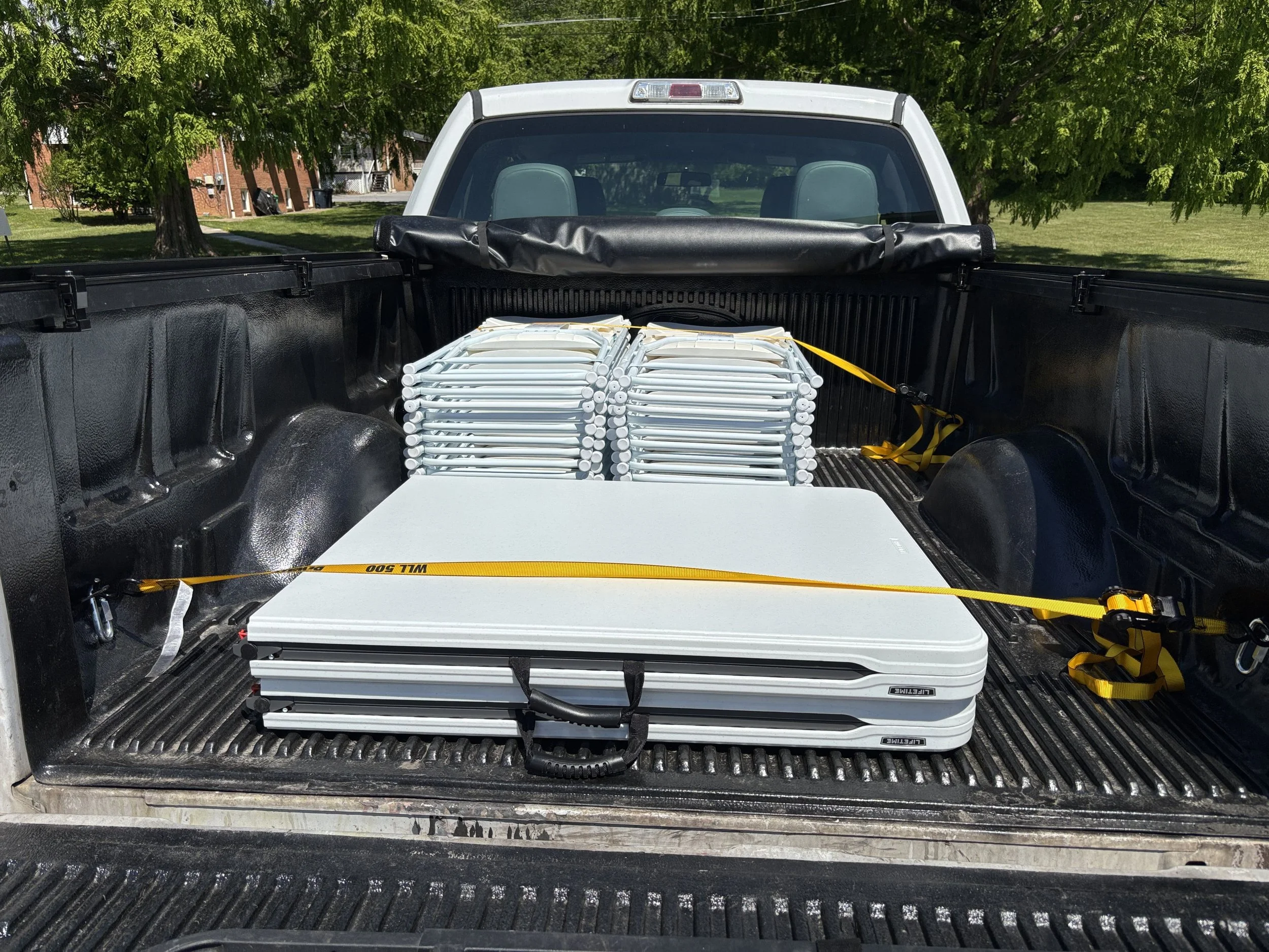 Back of a black pickup truck with a white folding table and a large bundle of white metal or plastic poles secured with yellow straps in the truck bed, with trees and buildings in the background.