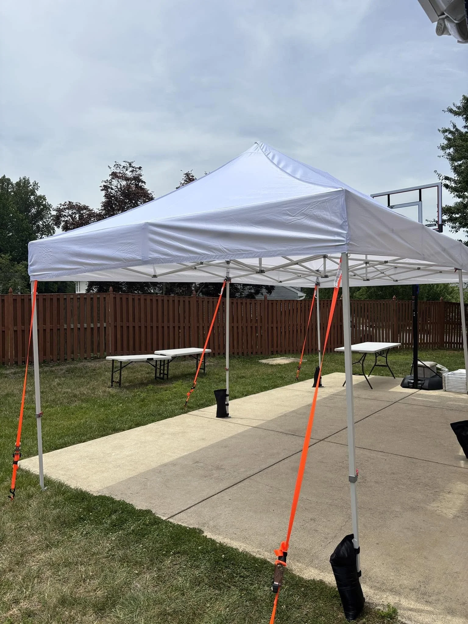 A white outdoor canopy tent set up on a grassy area with a concrete patio, with outdoor tables and a basketball hoop in the background, under a cloudy sky.