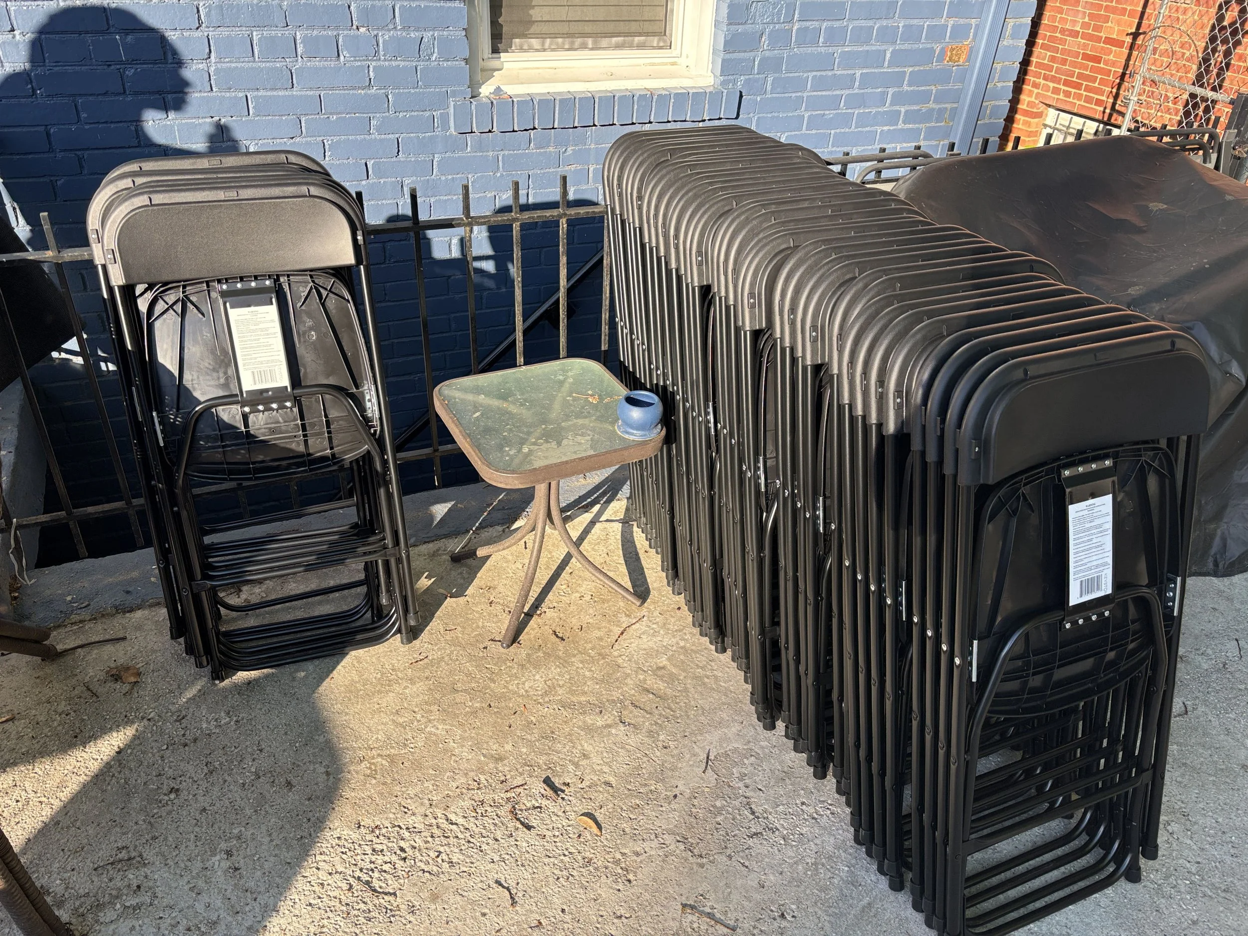 Stacked black metal folding chairs next to a small round table with a blue and black vase on top, placed outdoors on a concrete pavement in front of a blue brick wall and a window.