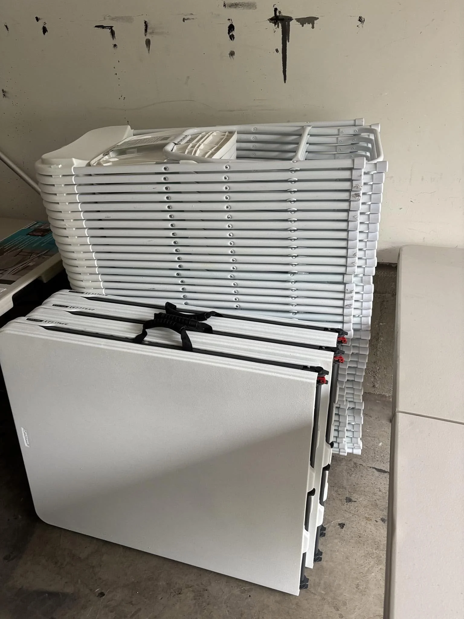 Stack of white folding tables leaning against a wall, with a small part of a white piece of furniture visible on the right side.