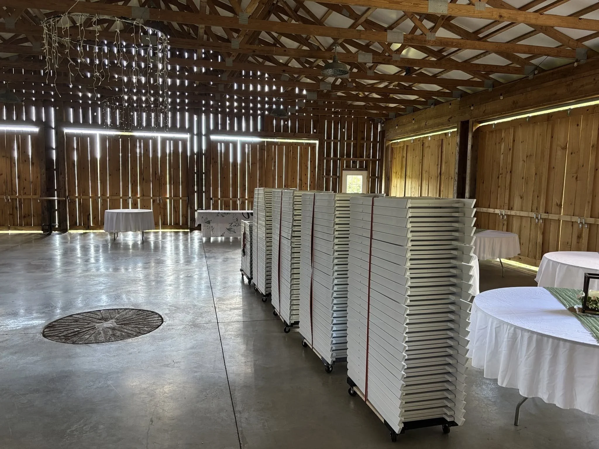 Inside a rustic barn with wooden walls and ceiling, decorated with string lights. There are stacks of white chairs on carts, and tablecloths with decorations on some round tables.