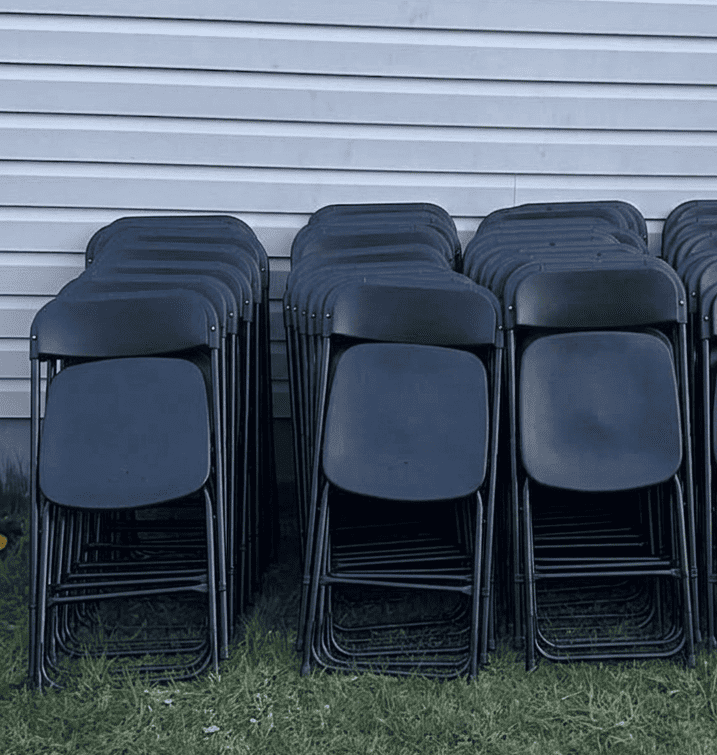 Multiple stacked black folding chairs outside against a white siding wall.