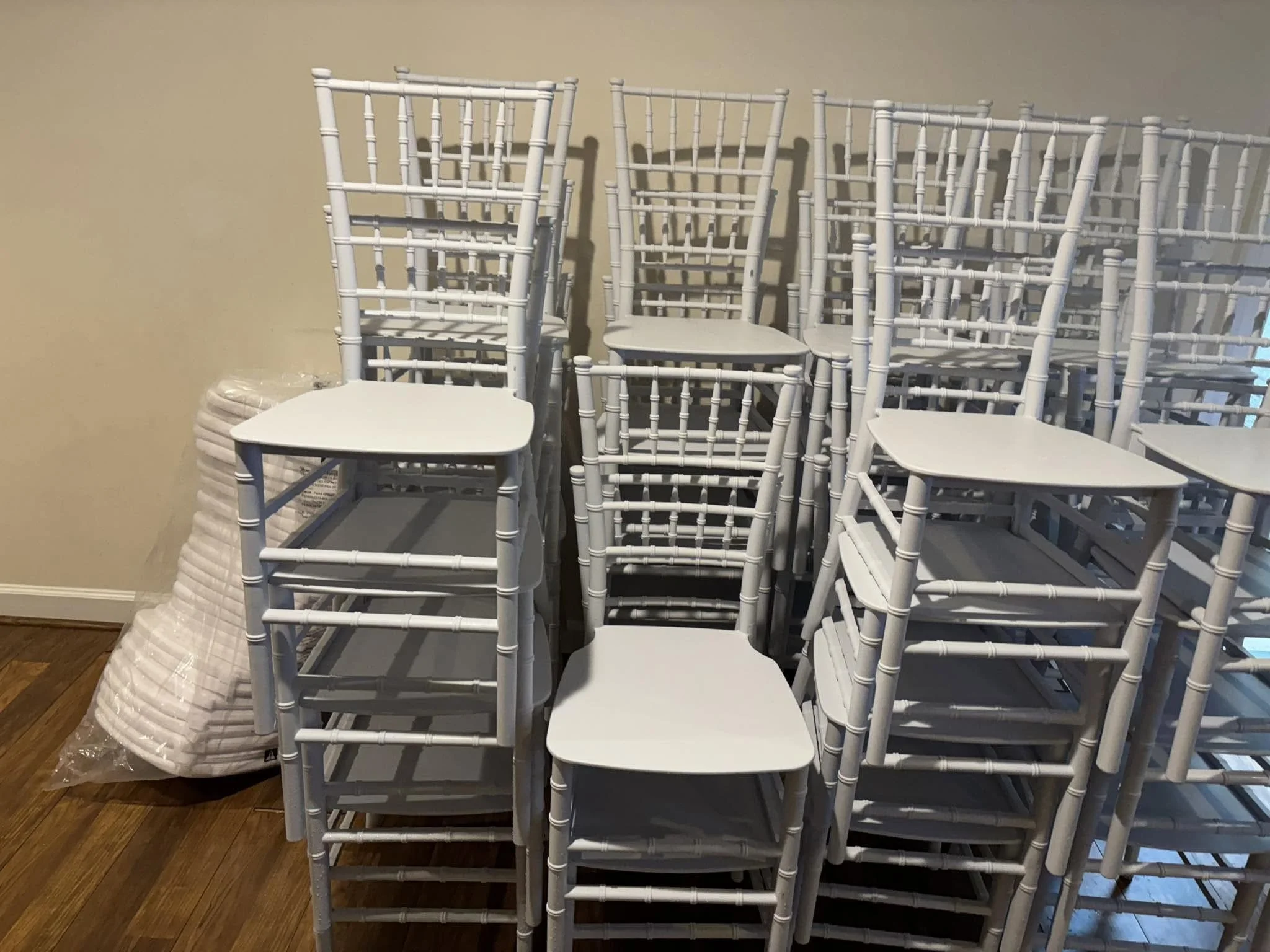 Stack of white folding chairs with a few chairs stacked to the side, placed against a beige wall and on a hardwood floor.