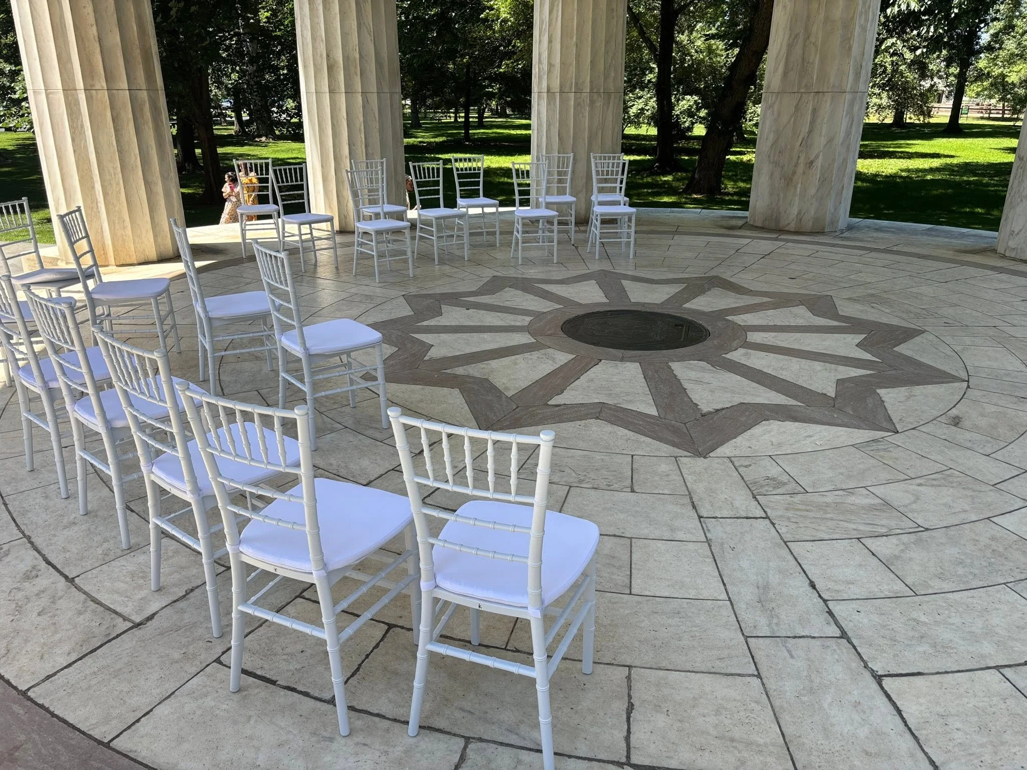 White chairs arranged in a circle inside a marble pavilion with columns, with a green park visible in the background.