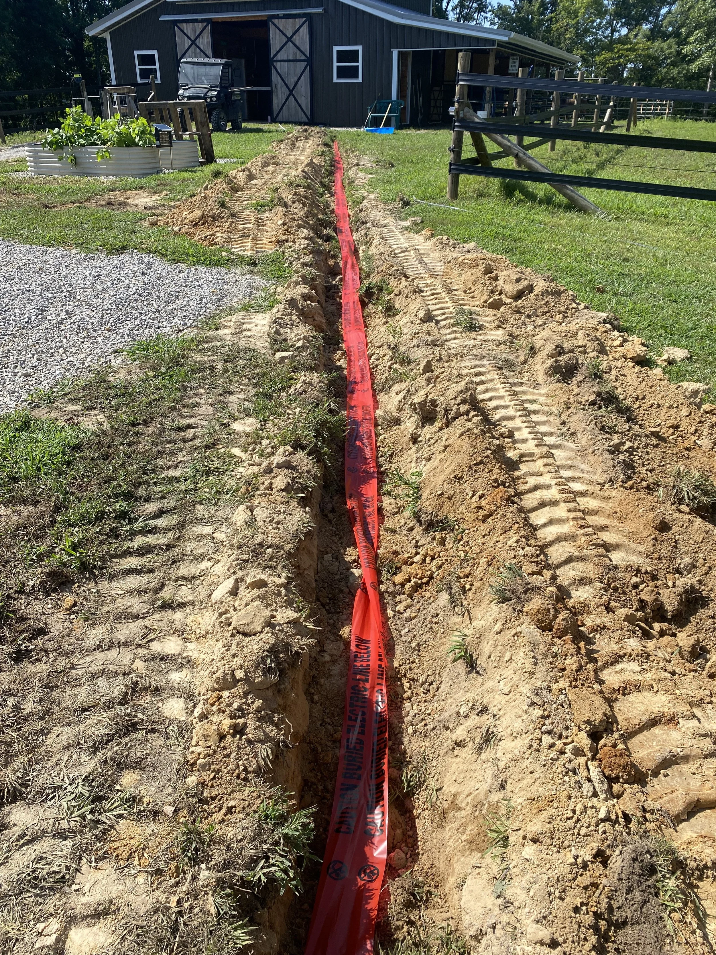 A construction site with a long trench dug in the ground, marked by red caution tape, next to a grassy yard and a barn with farm equipment in the background.